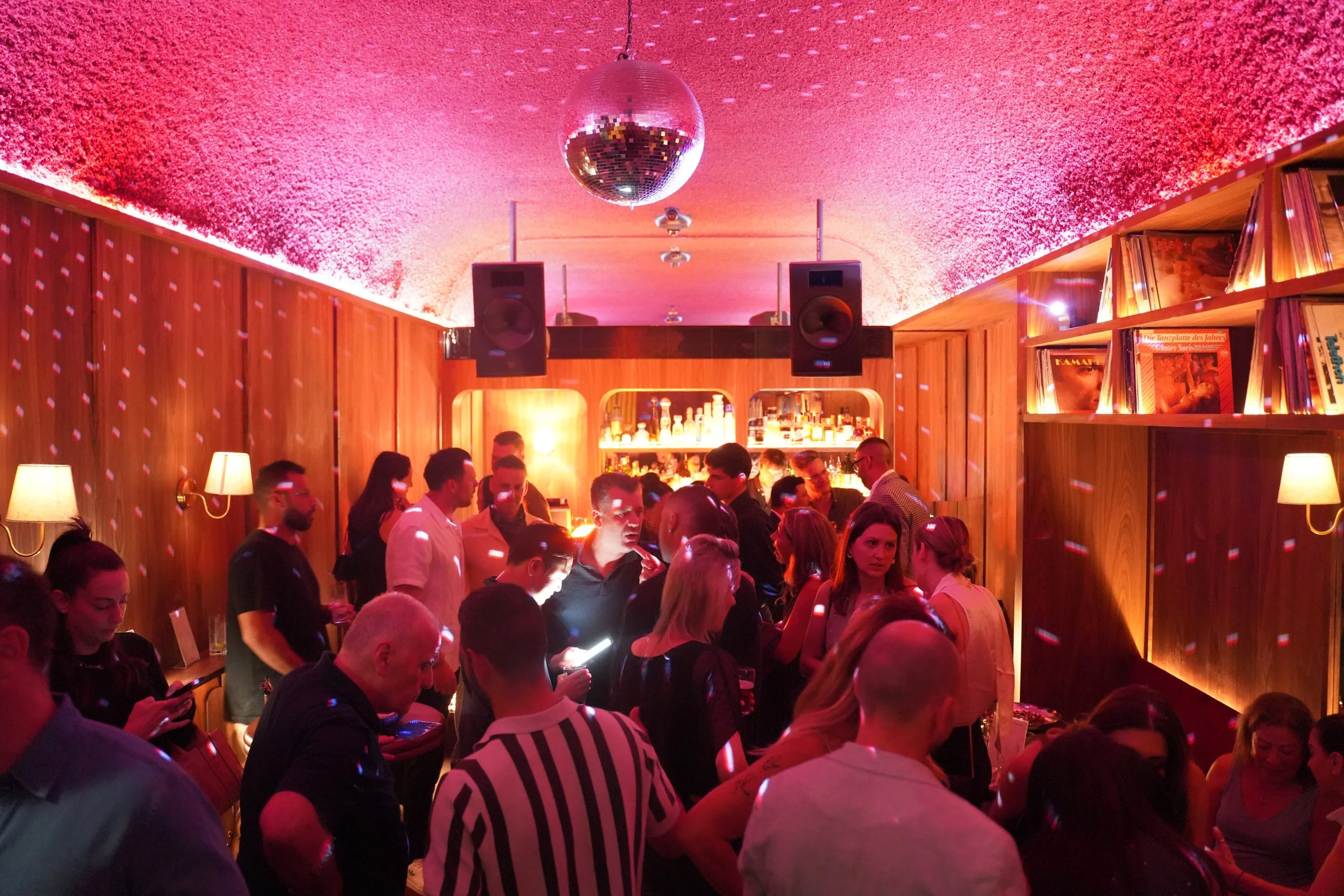 A DJ controller on a table in a dimly lit room with a red ceiling, disco balls, and wall sconces, suggesting a nightclub or party setting.