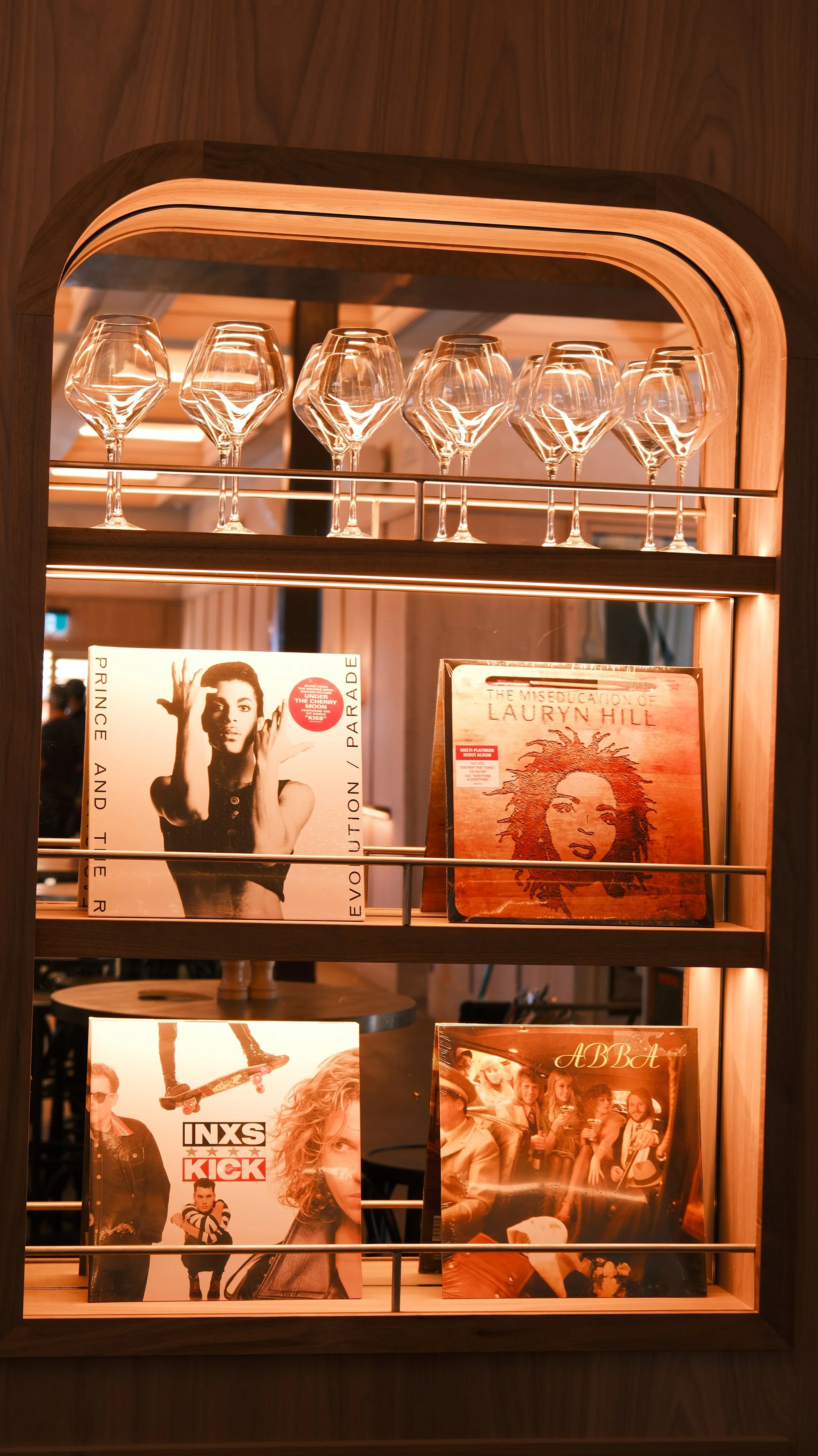 A wooden display shelf with wine glasses on top and vinyl records on the lower shelves, with album covers featuring Prince, Lauryn Hill, INXS, and ABBA visible.