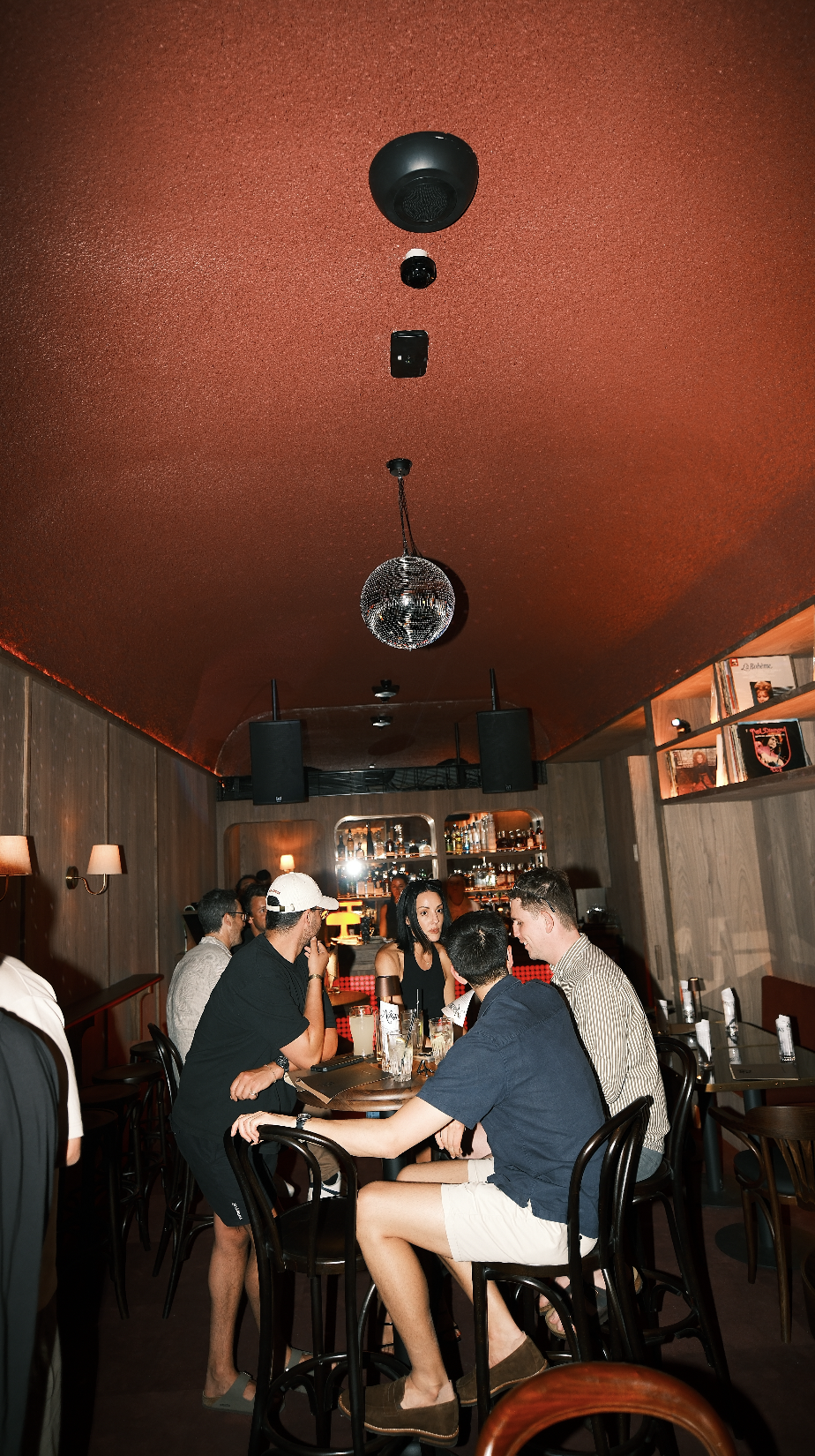 People sitting and talking at a bar or restaurant with wooden walls, shelves of bottles in the background, and a disco ball hanging from the ceiling.