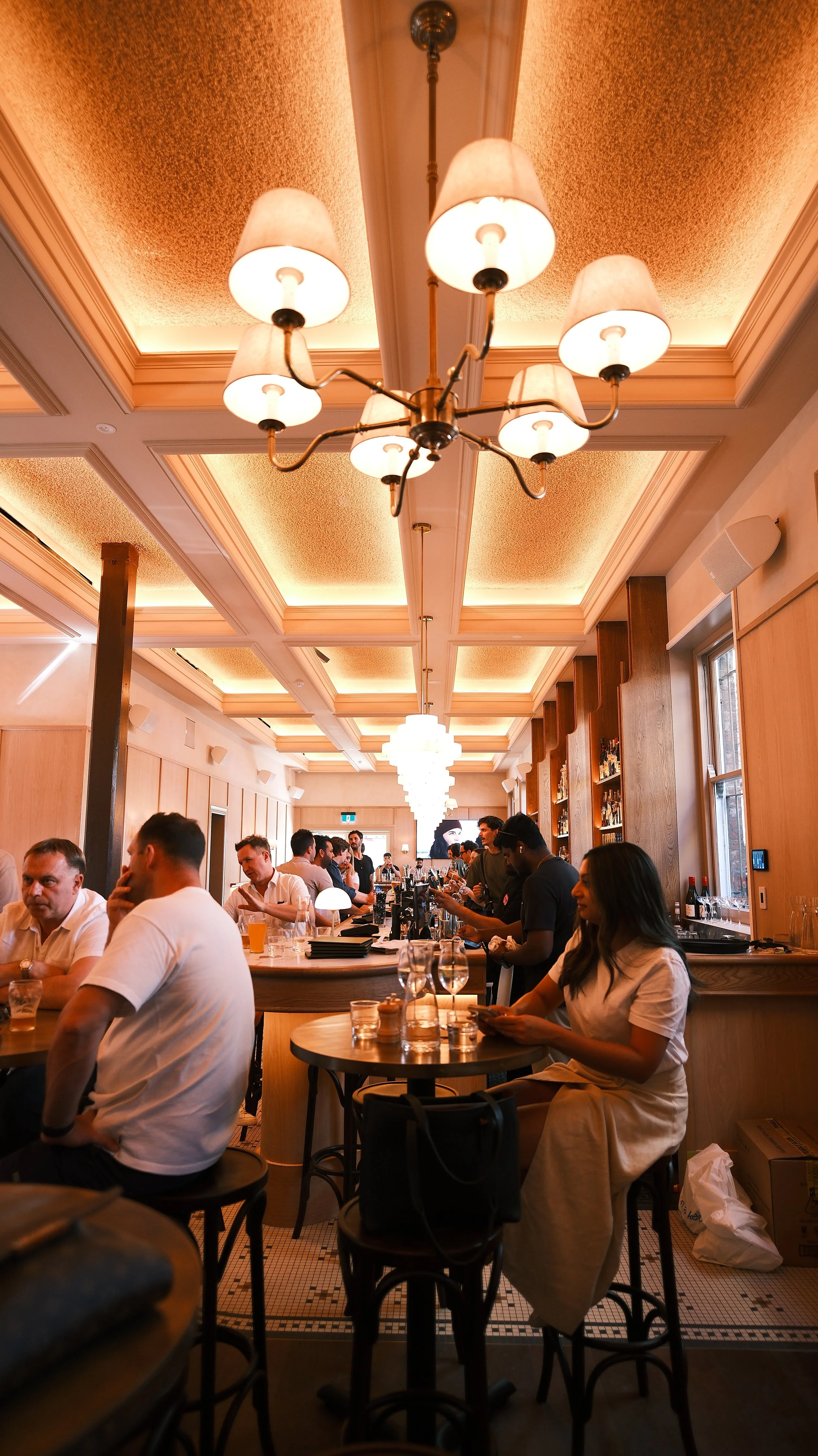 A busy bar inside a restaurant with warm lighting, pendant chandeliers, and a group of people socializing and enjoying drinks.
