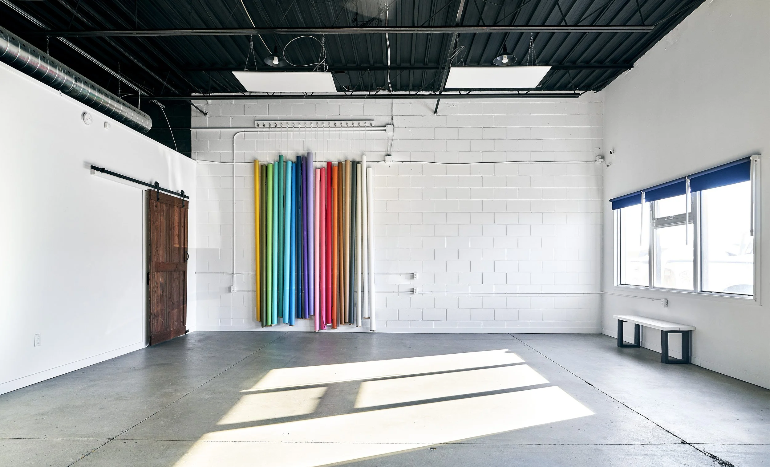 An empty artphotography studio with a white brick wall, large colorful seamless backdrops, a wooden barn door, large windows with blue blinds, and a small white bench near the windows.