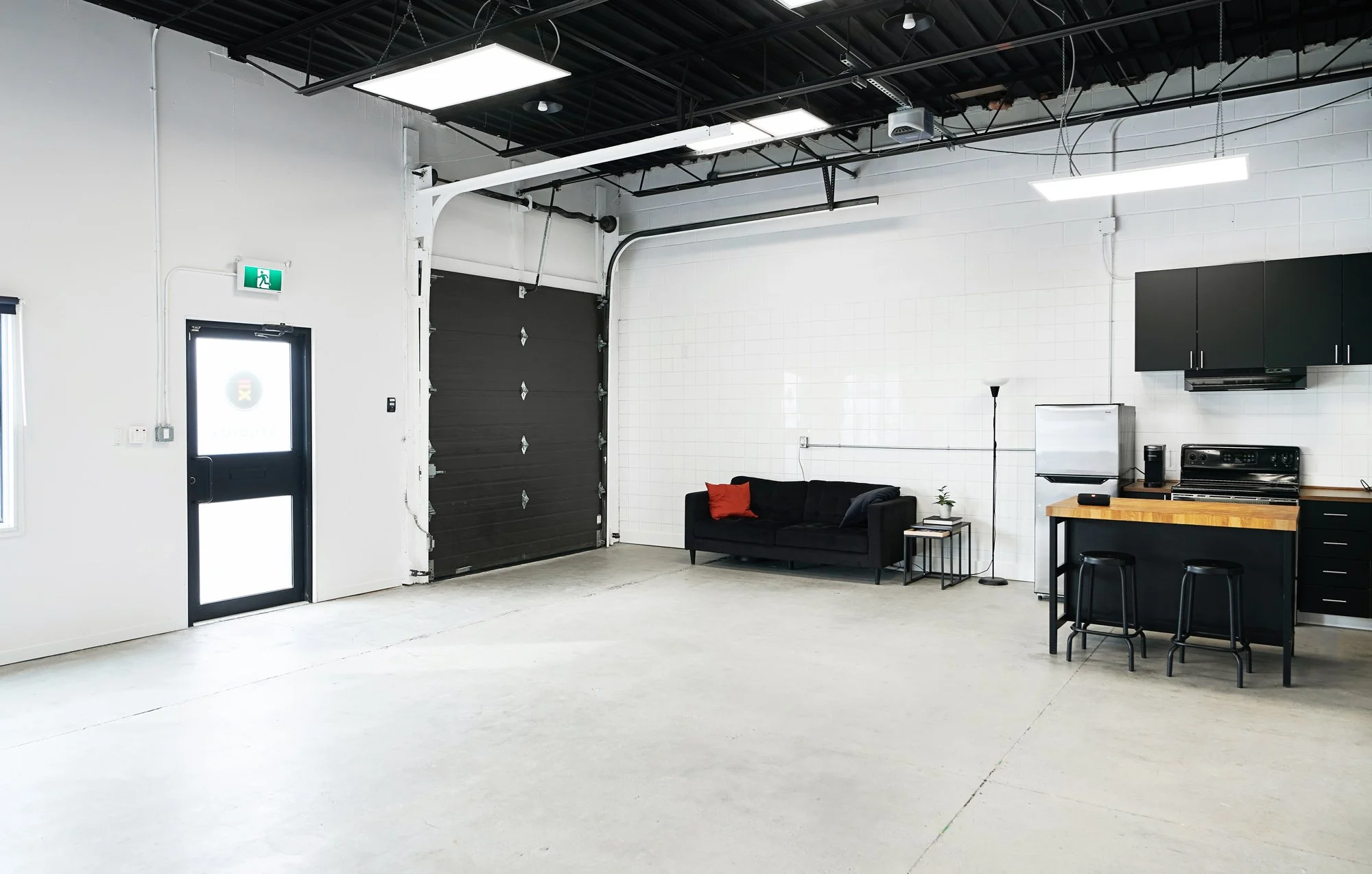 Spacious industrial-style interior with white walls, concrete floor, black leather sofa with red and black pillows, small side table with plant, black floor lamp, kitchenette with black cabinets, stainless steel mini refrigerator, black stove and mic