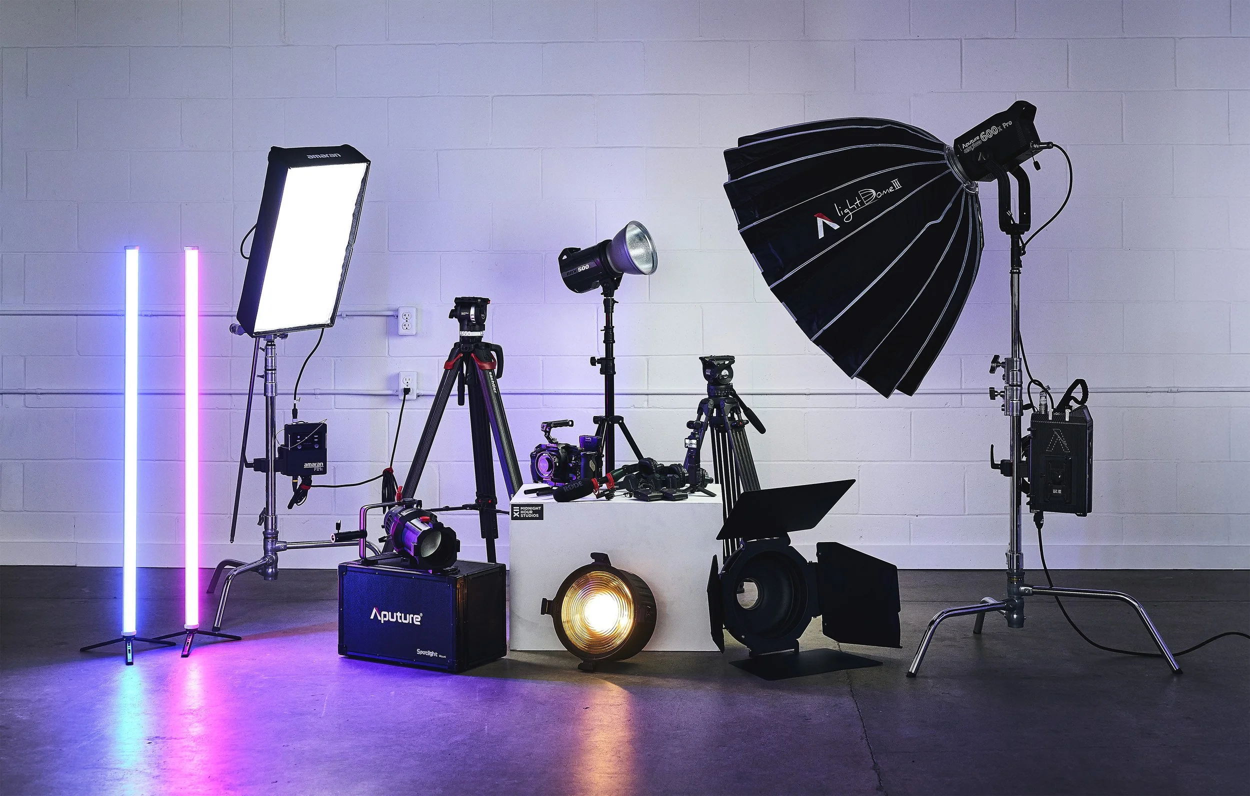 A professional photography or video shoot setup with various lighting equipment, tripods, and reflectors arranged indoors against a white brick wall.