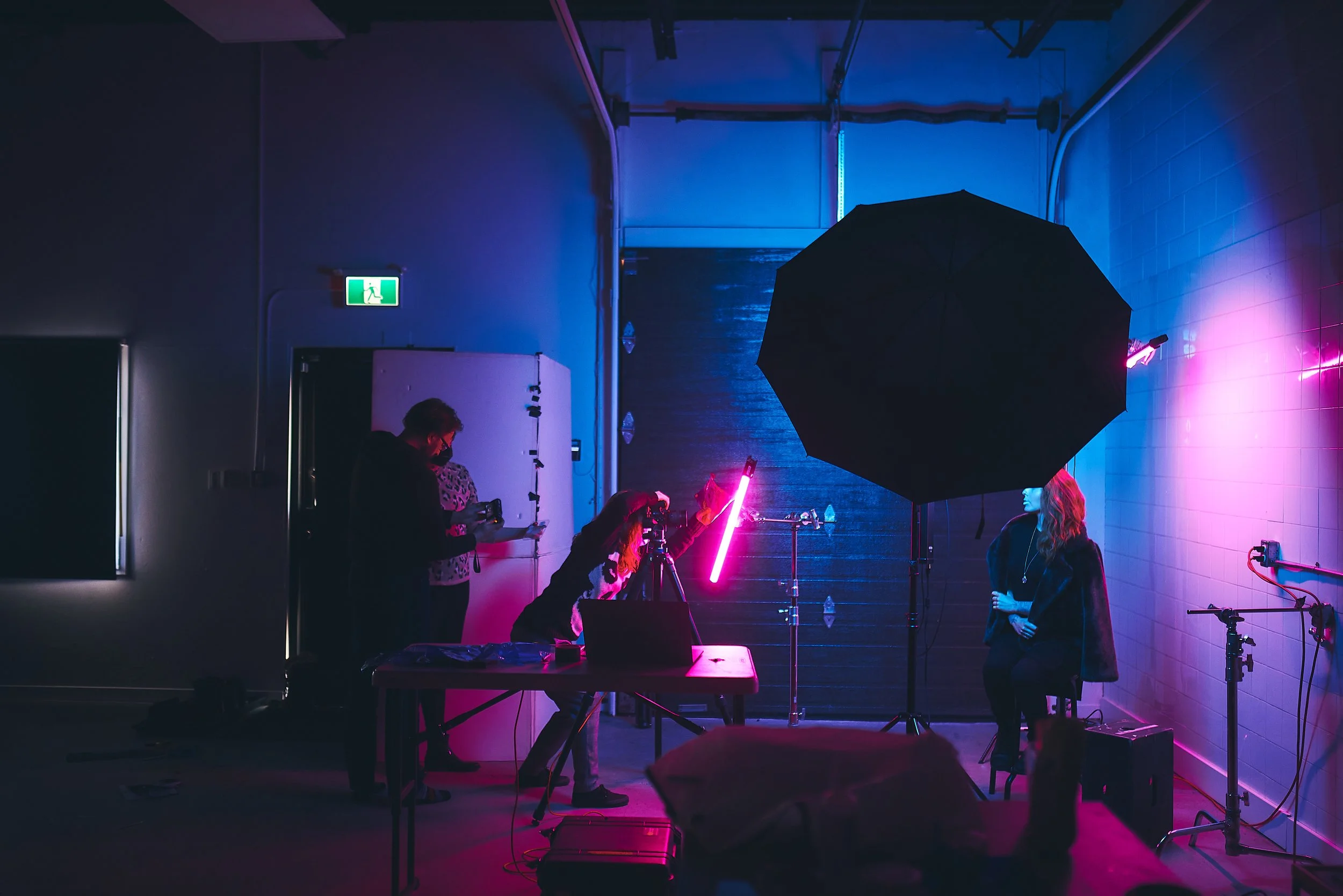People working on set with photography equipment, neon lights, and a black umbrella in a dimly lit studio