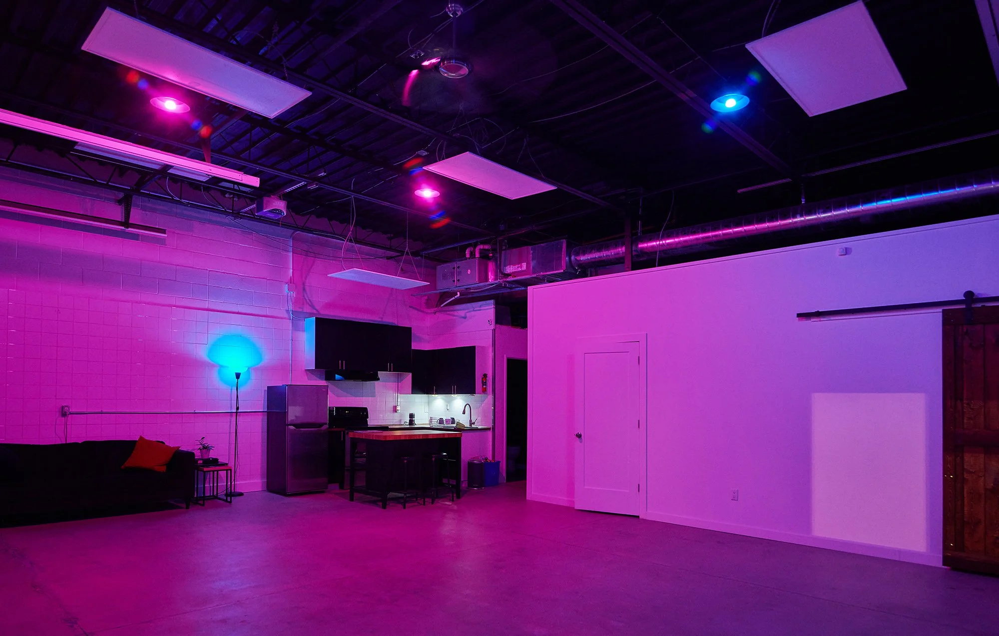 An indoor space illuminated with pink and purple lighting, featuring a black sofa with orange and red cushions, a nightstand, a floor lamp with blue light, a small kitchenette with a refrigerator, countertop, sink, and black cabinets, and a white wal