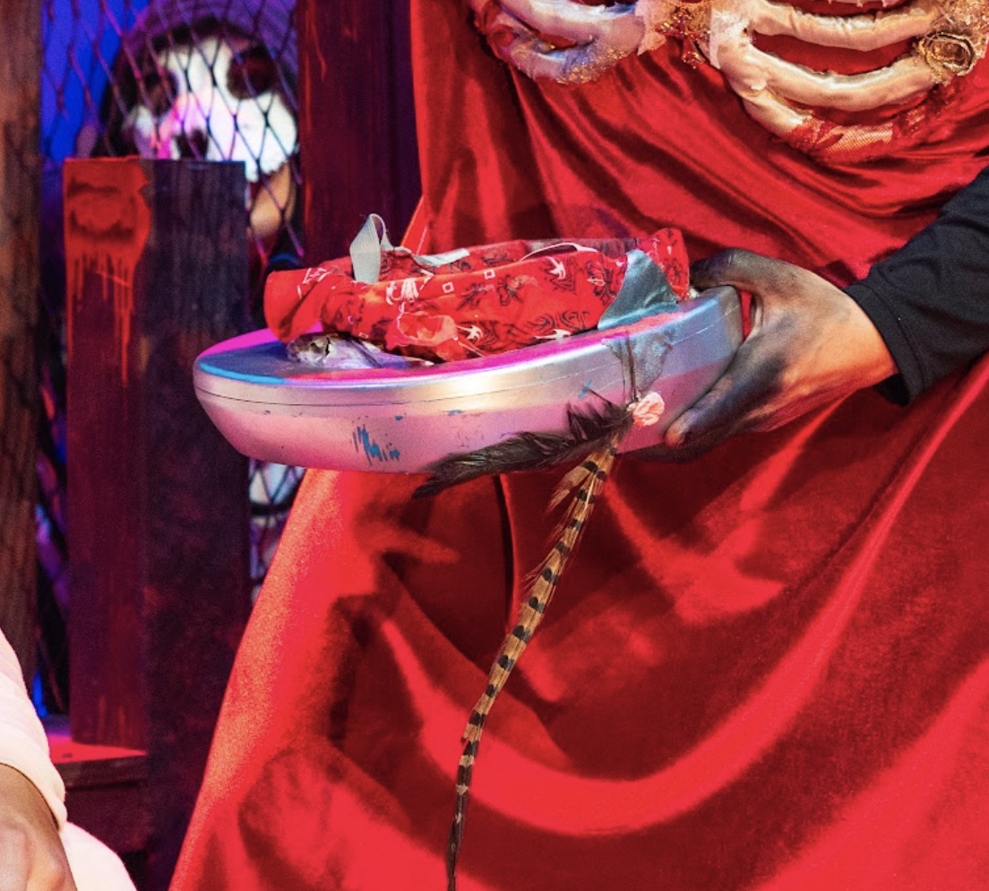 Bed pan helmet on stage. Plastic bedpan painted silver mounted on fosshape cap. Cap wrapped in a red bandana to hide it.