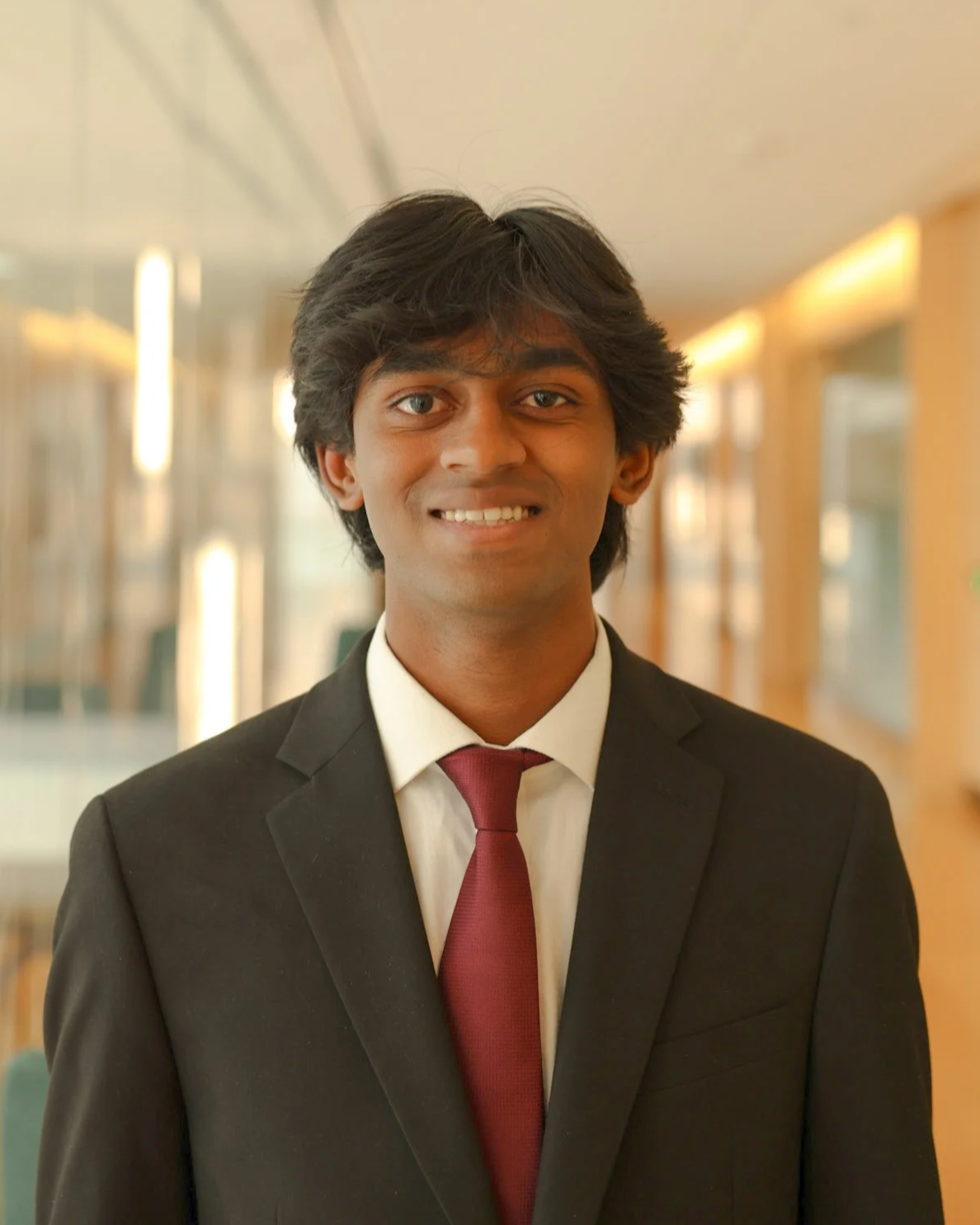Young man in black suit, white shirt, and red tie standing in a well-lit corridor with glass walls and yellow framing.