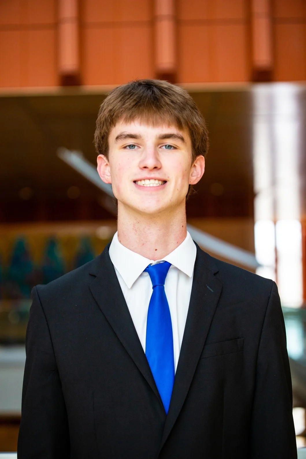 A young man wearing a black suit, white shirt, and blue tie, standing indoors with blurred background of wooden and metallic elements.