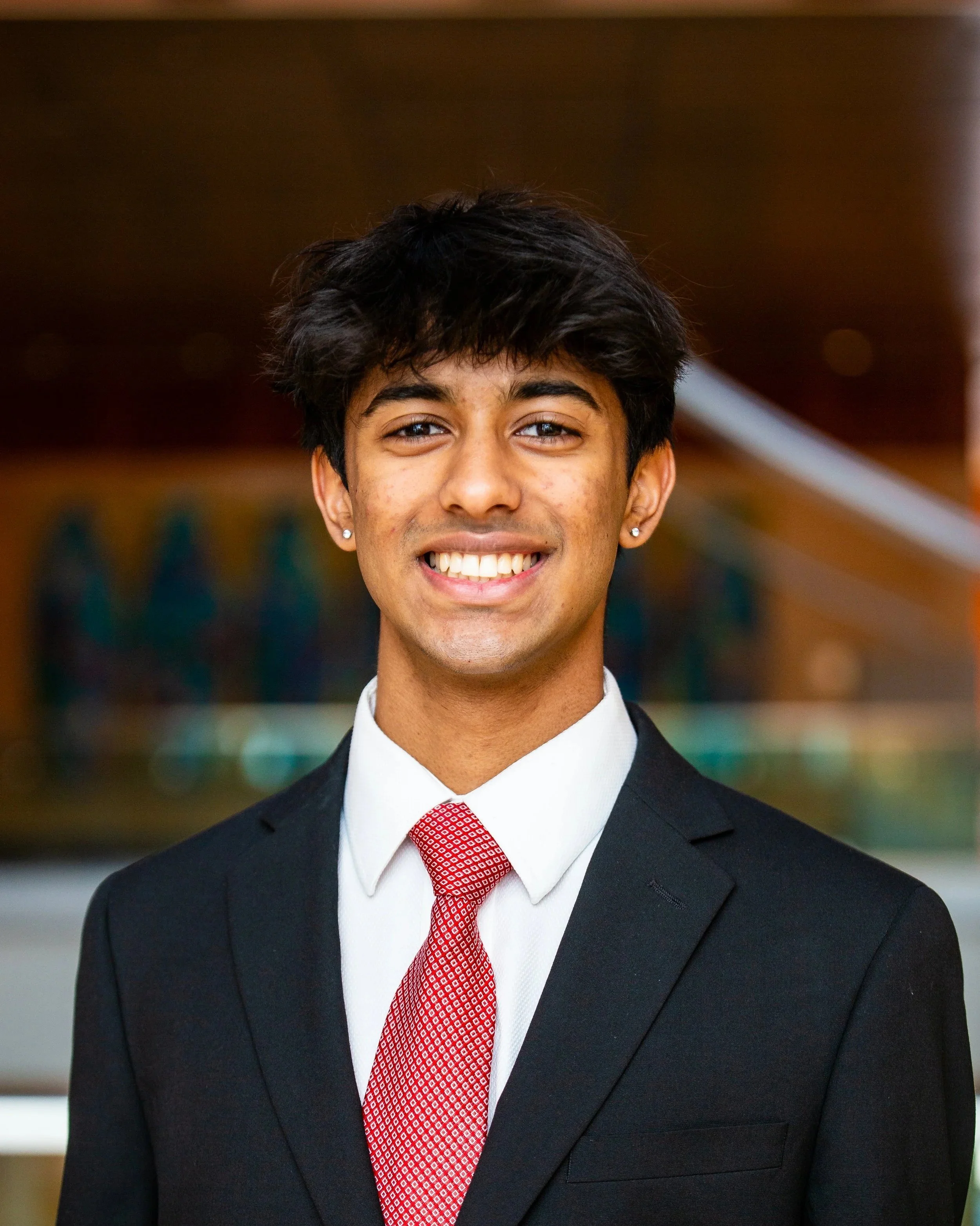 A young man smiling, dressed in a black suit, white shirt, and red patterned tie, with short dark hair and earrings.
