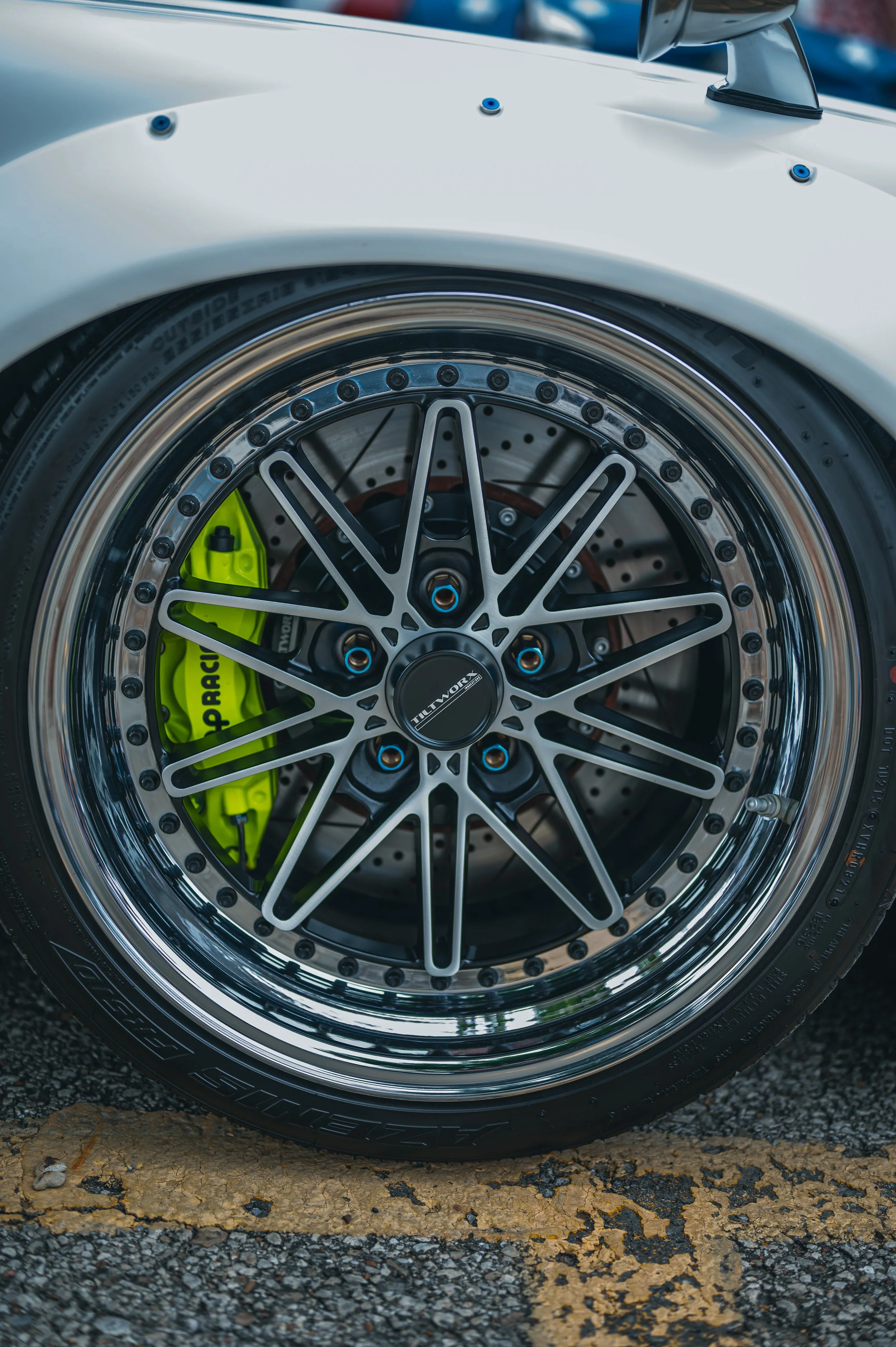 Close-up of a car wheel with a detailed alloy rim, bright green brake caliper, and a tire on pavement with peeling yellow paint.