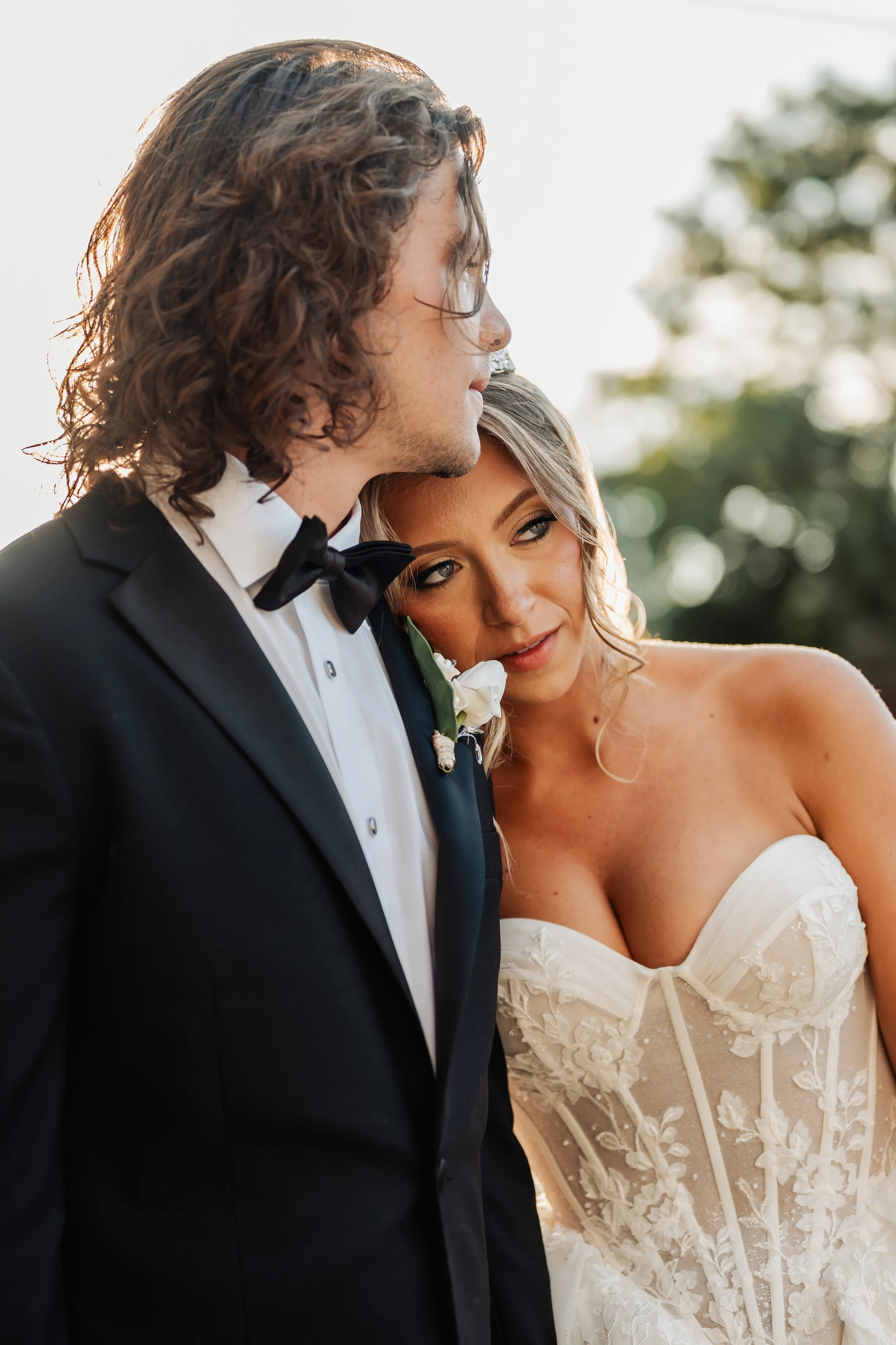 A bride and groom dressed in wedding attire, standing close to each other outdoors during sunset, with the groom wearing a black tuxedo and the bride wearing a white lace wedding gown.