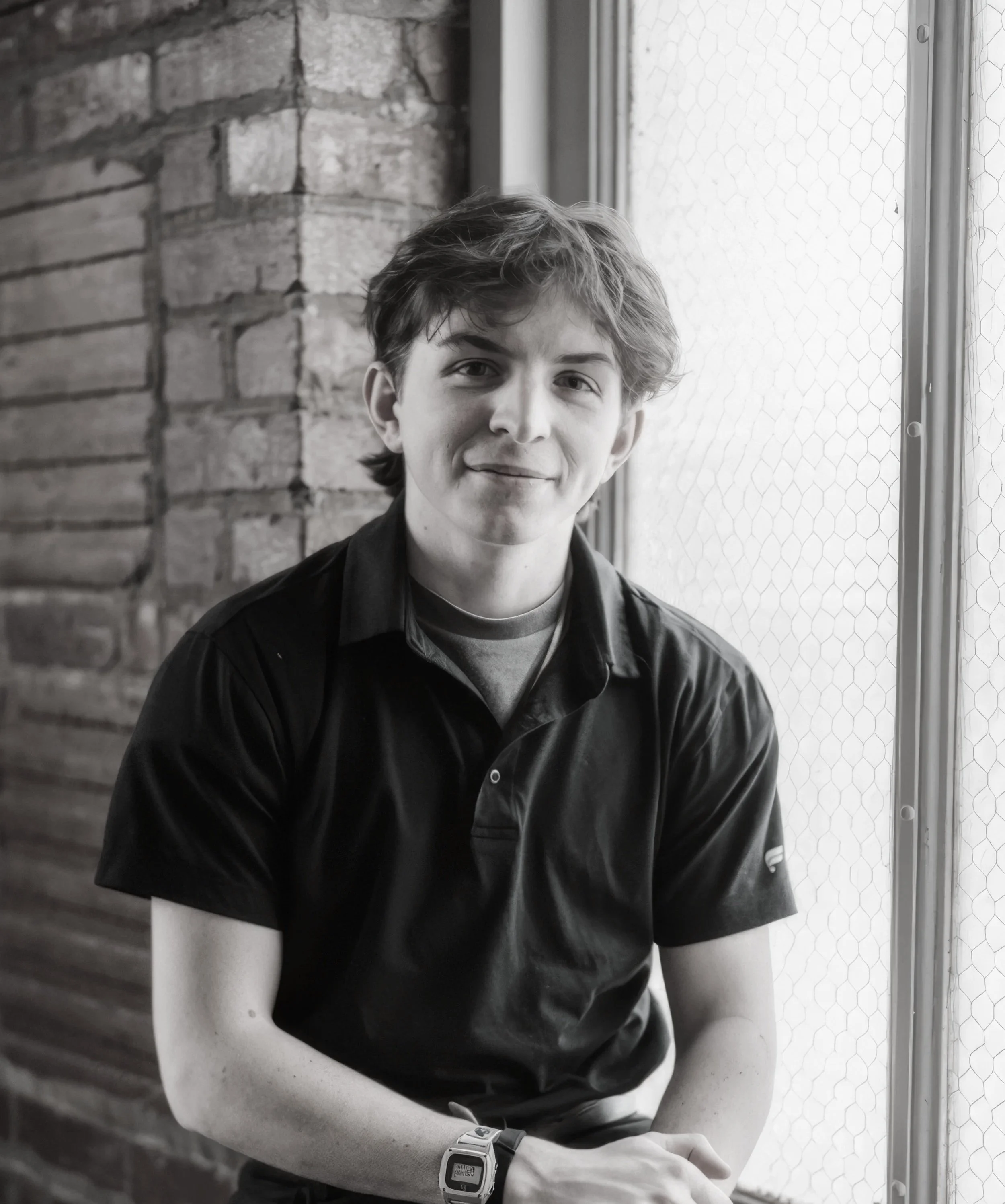 A young man with wavy hair sitting by a window with a brick wall behind him, smiling at the camera.