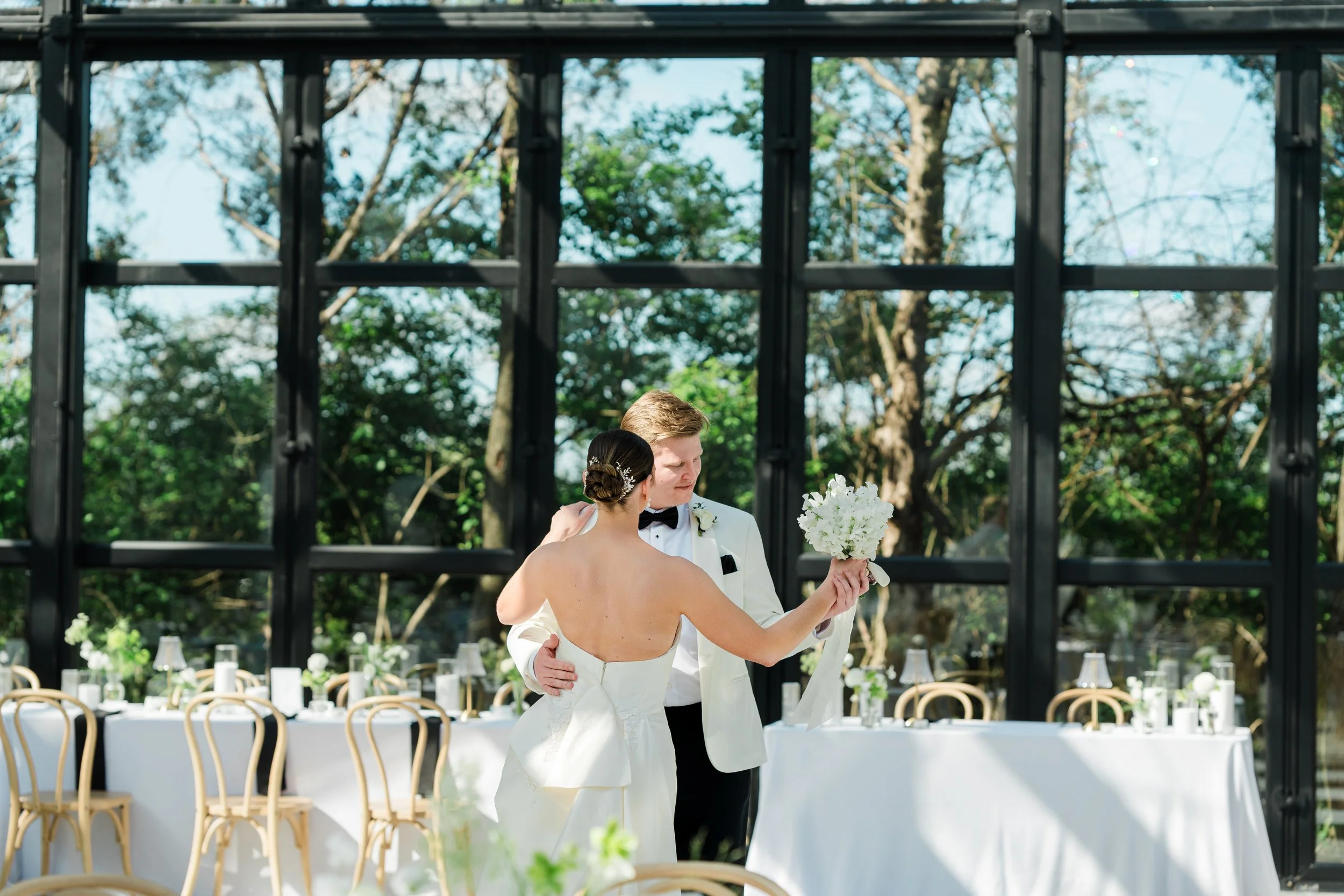 A wedding couple, with the bride holding a bouquet, sharing a dance outdoors at a reception area decorated with white tablecloths and chairs, under a structure with a black metal framework and surrounded by trees.