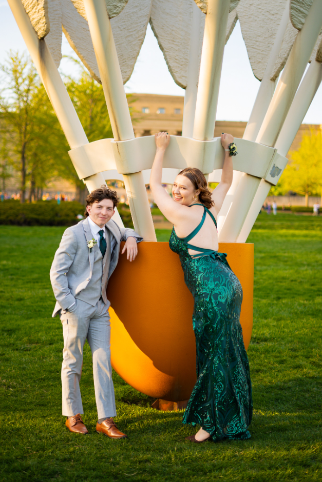 A young man in a light gray suit and a young woman in an emerald green gown with cutouts poses near a hot air balloon sculpture in a park during daylight. The woman is smiling and holding onto the sculpture, while the man is leaning against it with one hand in his pocket.