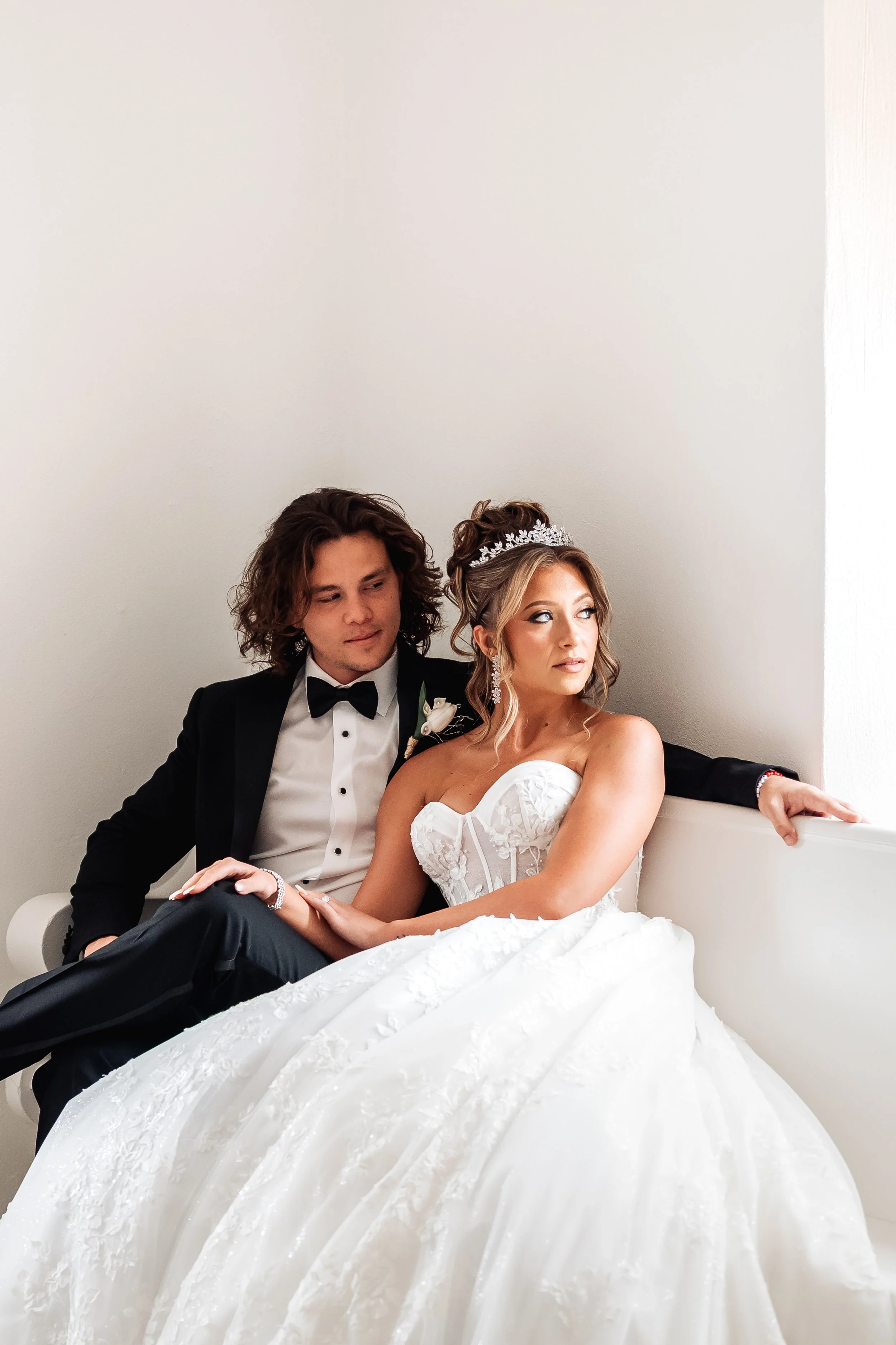 A bride and groom sit close together on a white sofa, with the groom in a black tuxedo and the bride in a white wedding gown and tiara, against a plain white wall background.