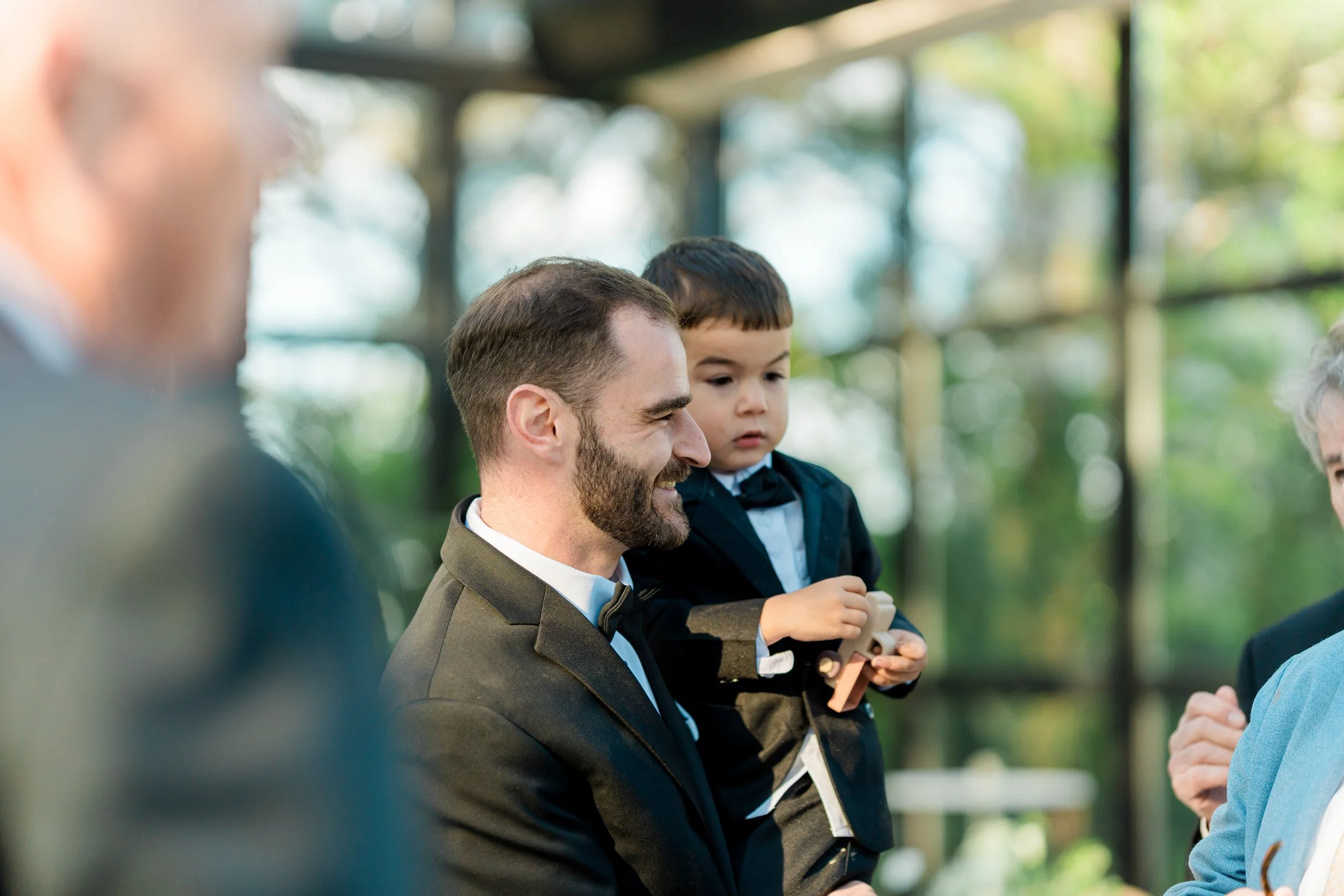 A man in a suit and tie smiling as he talks to a woman outside, while a young boy in a blazer and bowtie holds a paper and looks on.