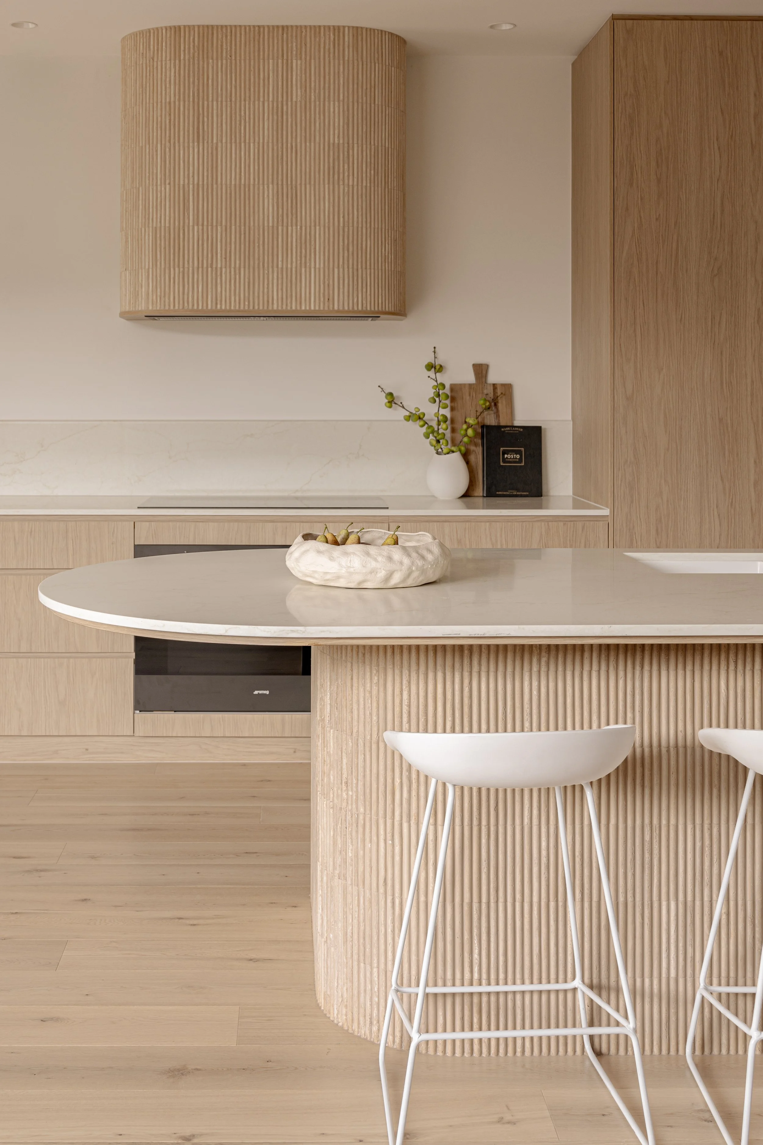 A minimalist kitchen with light wood cabinets, a white marble countertop, and white bar stools with metal legs. There is a decorative bowl with fruit on the island, and a vase with green berries, a cutting board, and a black box on the counter in the background.