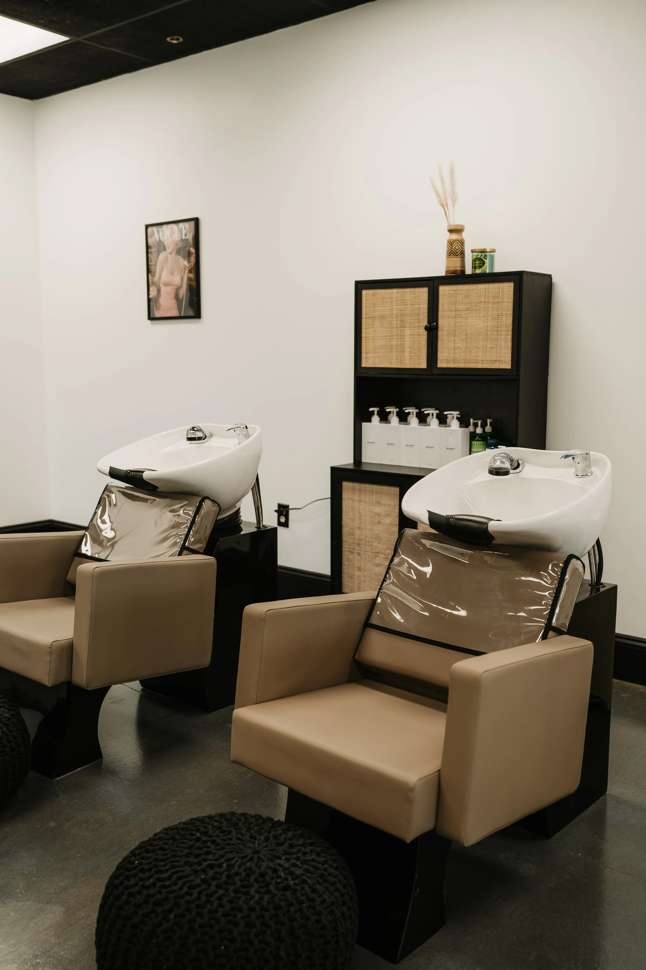 Two salon shampoo stations with beige chairs and white sinks, a black storage cabinet with bottles of hair products, a framed Vogue magazine cover on the wall, and a decorative vase with dried plants on top of the cabinet.