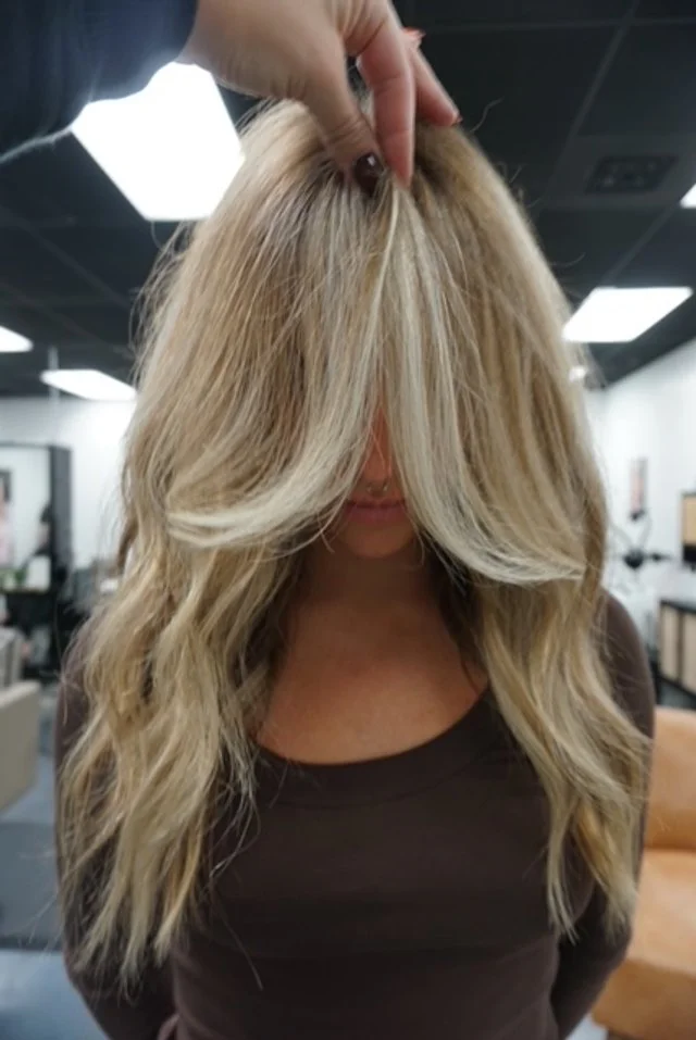 A woman with long blonde wavy hair having her roots touched up at a salon.