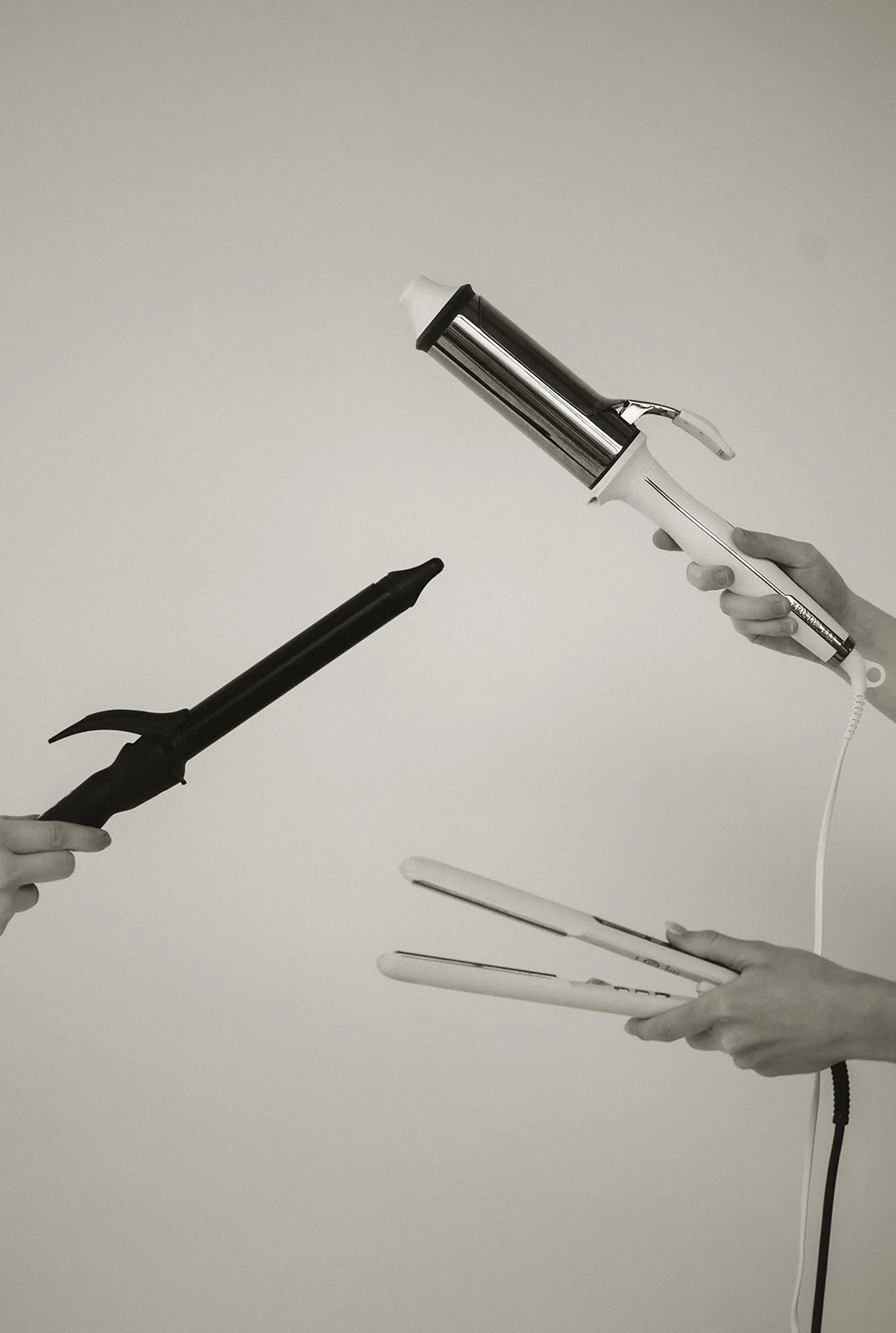 Three hands holding different styling tools: a curling iron, a curling wand, and a flat iron, against a plain background.