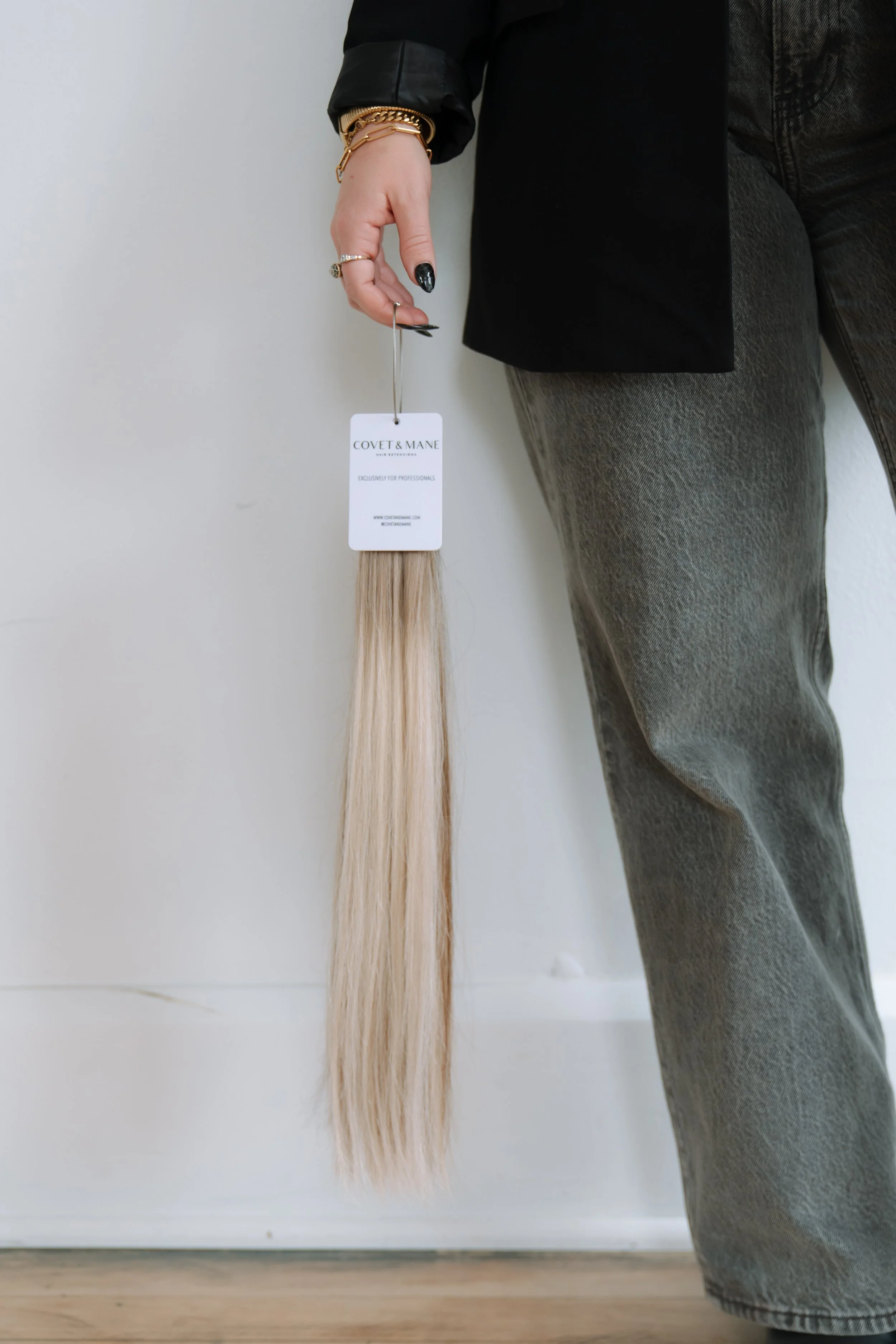 Person holding a hair extension sample labeled 'COVET & MANE' in front of a white wall, dressed in dark pants and a black jacket with jewelry.