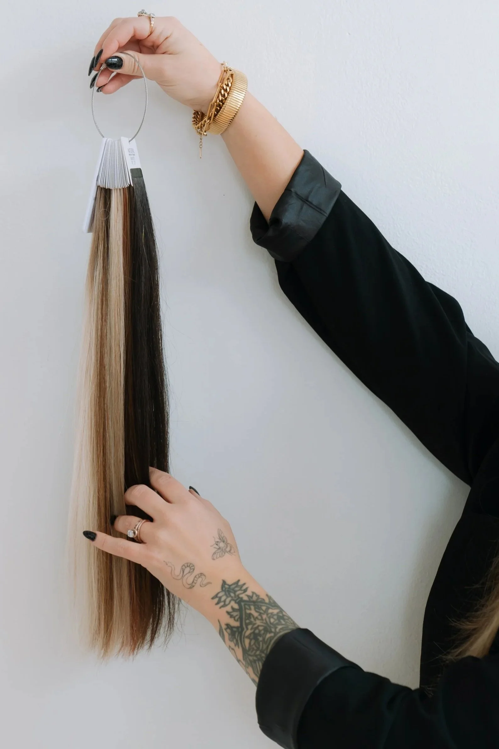 A person with tattooed hands holding a hair color swatch with strands of blonde, light brown, dark brown, and black hair, wearing a black long sleeve and gold jewelry.