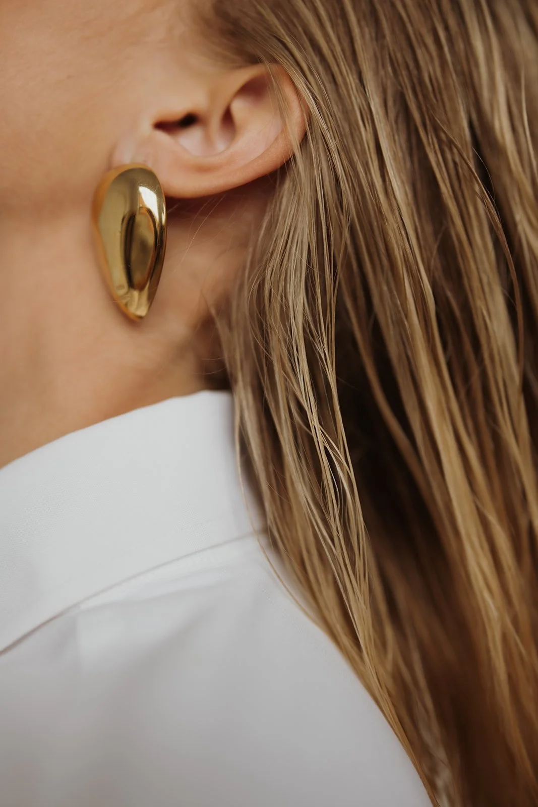 Close-up of a woman wearing a large, gold, irregularly-shaped earring and a white shirt, with long wavy hair.