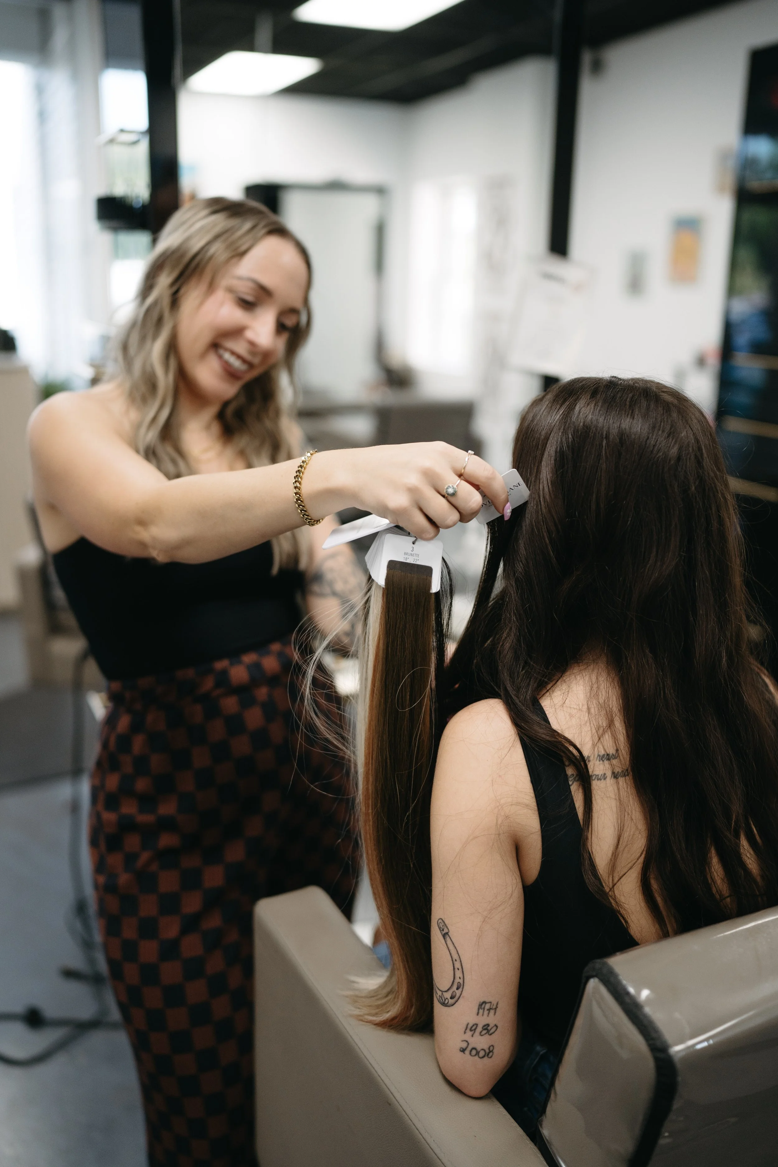 A woman with long blonde hair is smiling as she holds a sample of hair color swatch near a woman's hair, inside a hair salon.