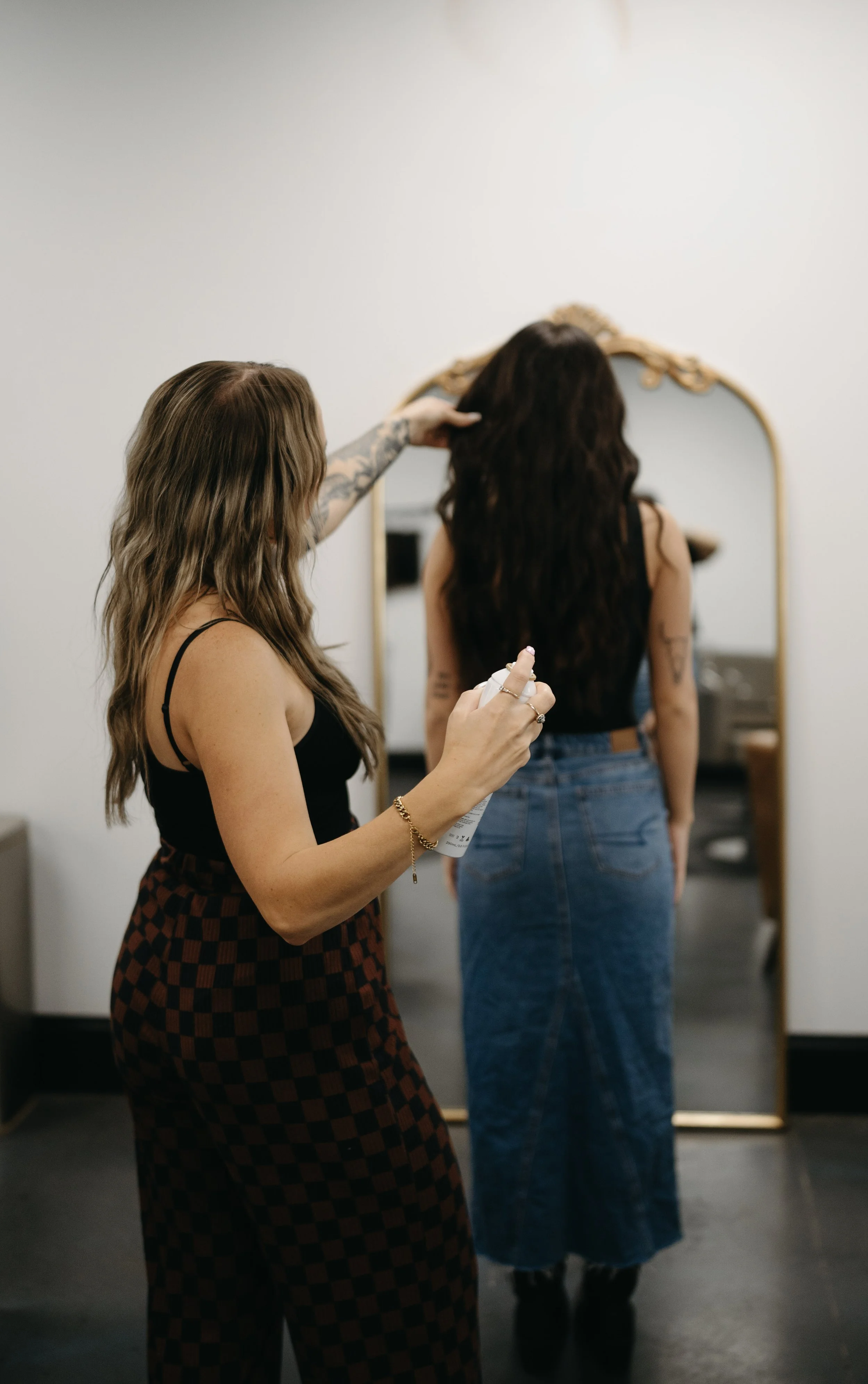 A woman with long, wavy, brown hair being styled by another woman with long, light brown hair, in front of a large, ornate gold-framed mirror inside a room.
