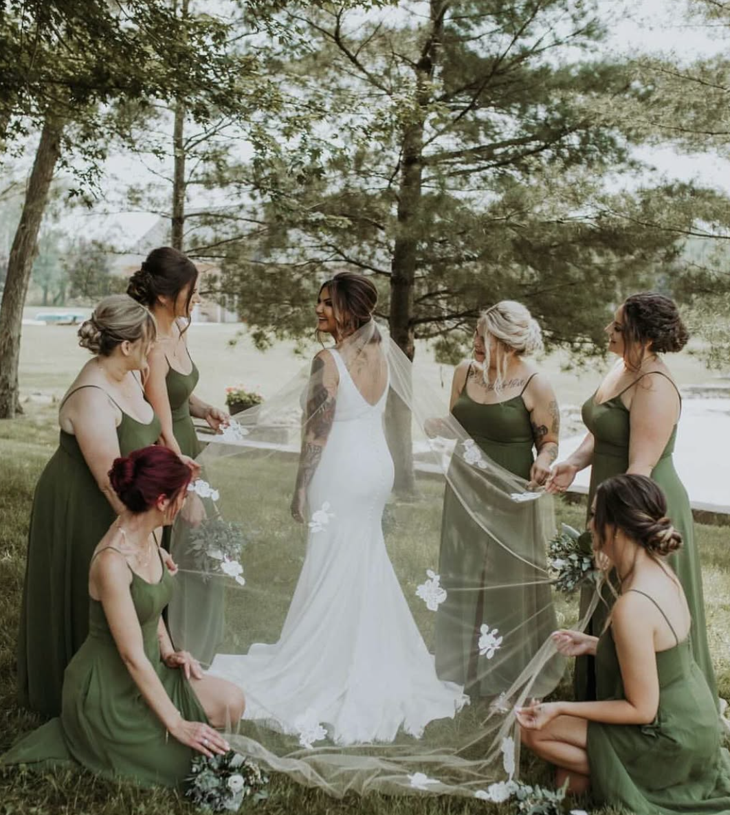 A bride surrounded by six bridesmaids outdoors near a lake, under trees, holding a sheer veil with floral appliqués.