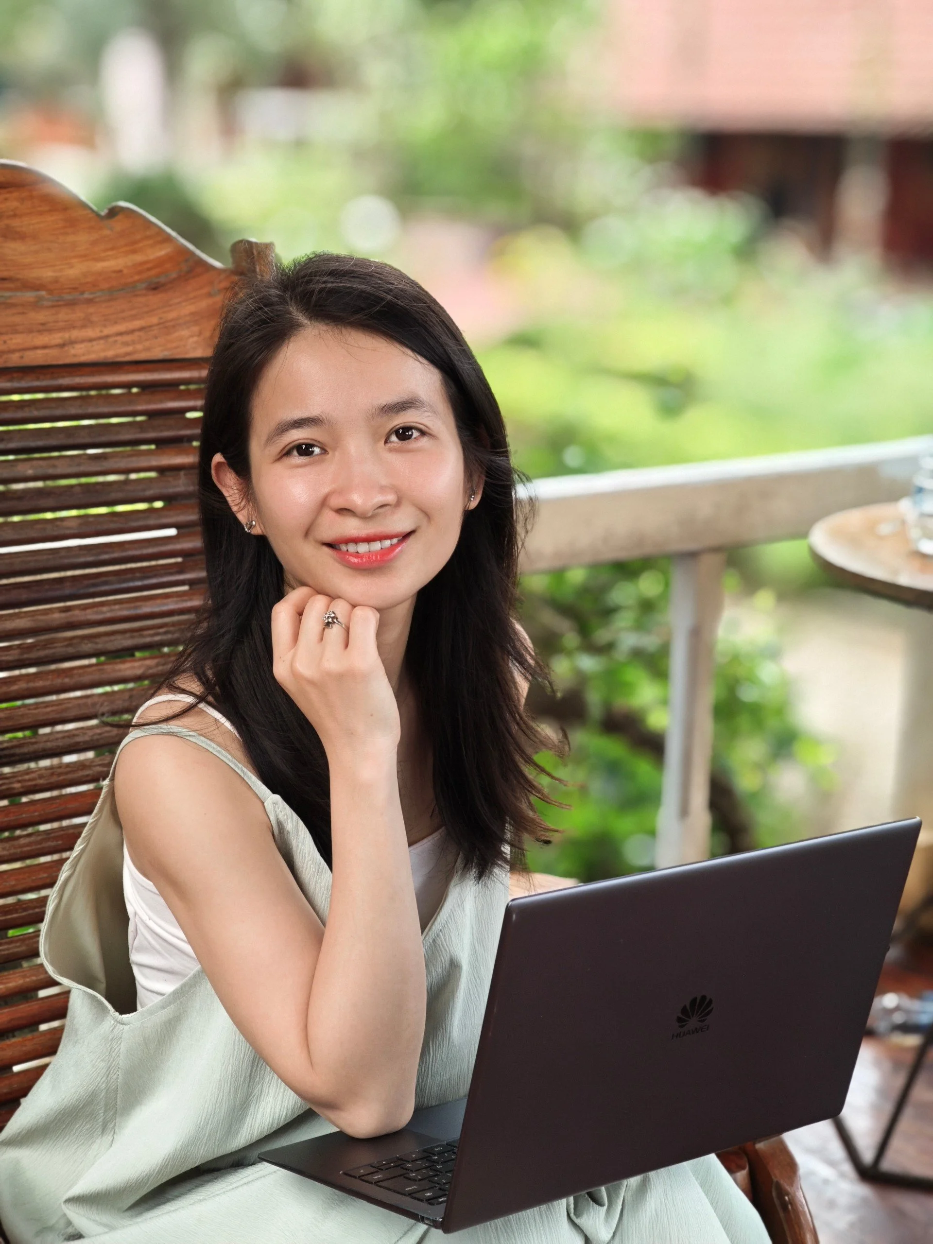 A young woman with long dark hair and earrings sitting outdoors at a wooden table with a laptop, smiling at the camera, with green trees blurred in the background.