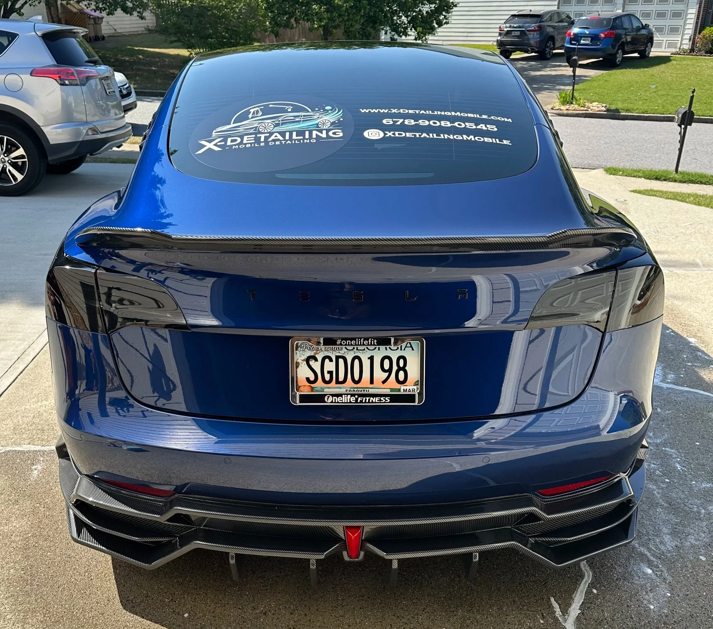 Rear view of a blue Tesla car with a carbon fiber spoiler and a custom license plate SGD0198, parked on a driveway with other vehicles and a neighborhood street in the background.