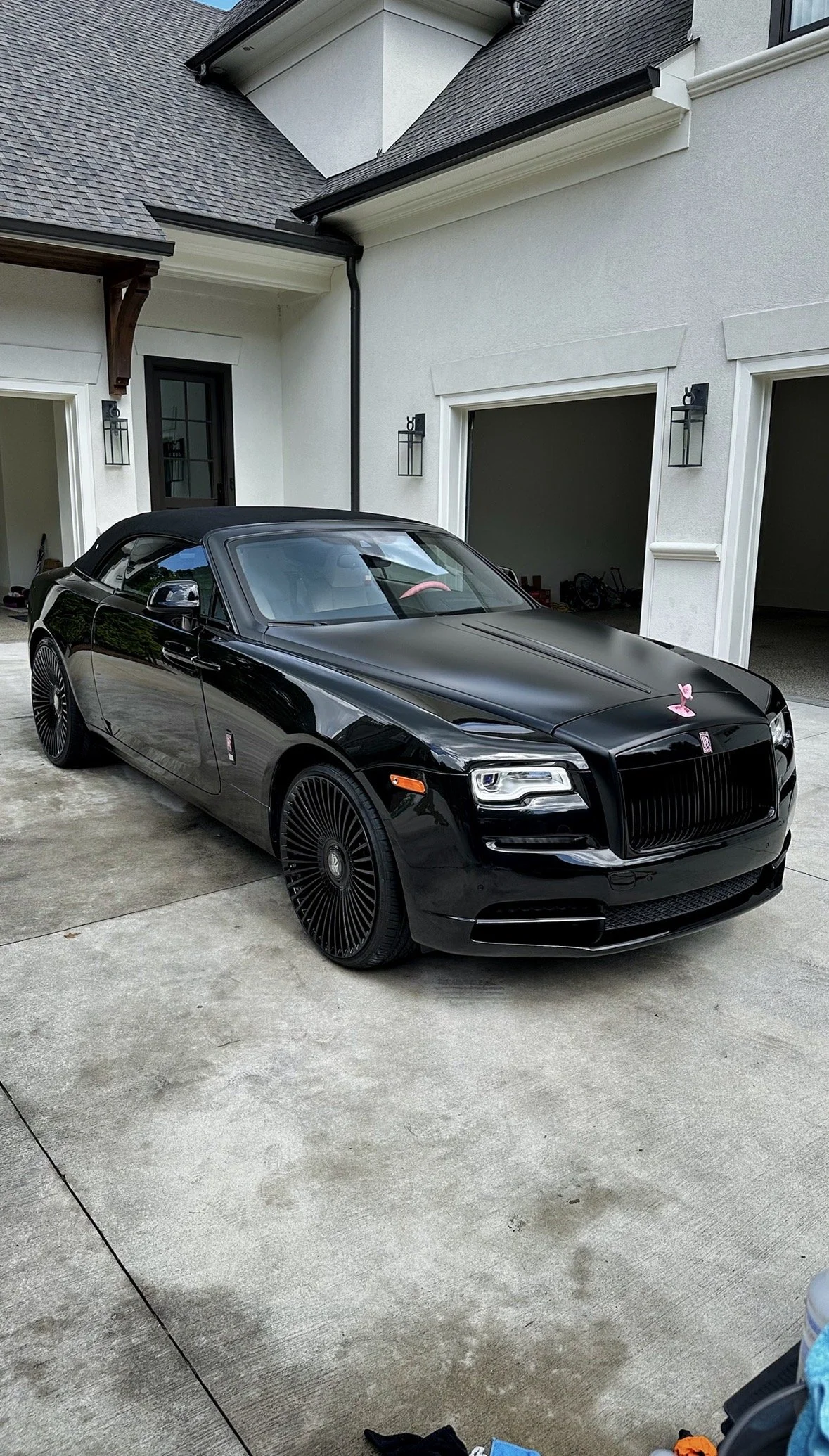 A black luxury convertible car parked in front of a white house with a garage, pink decorations on the hood, and black alloy wheels with intricate spoke designs.