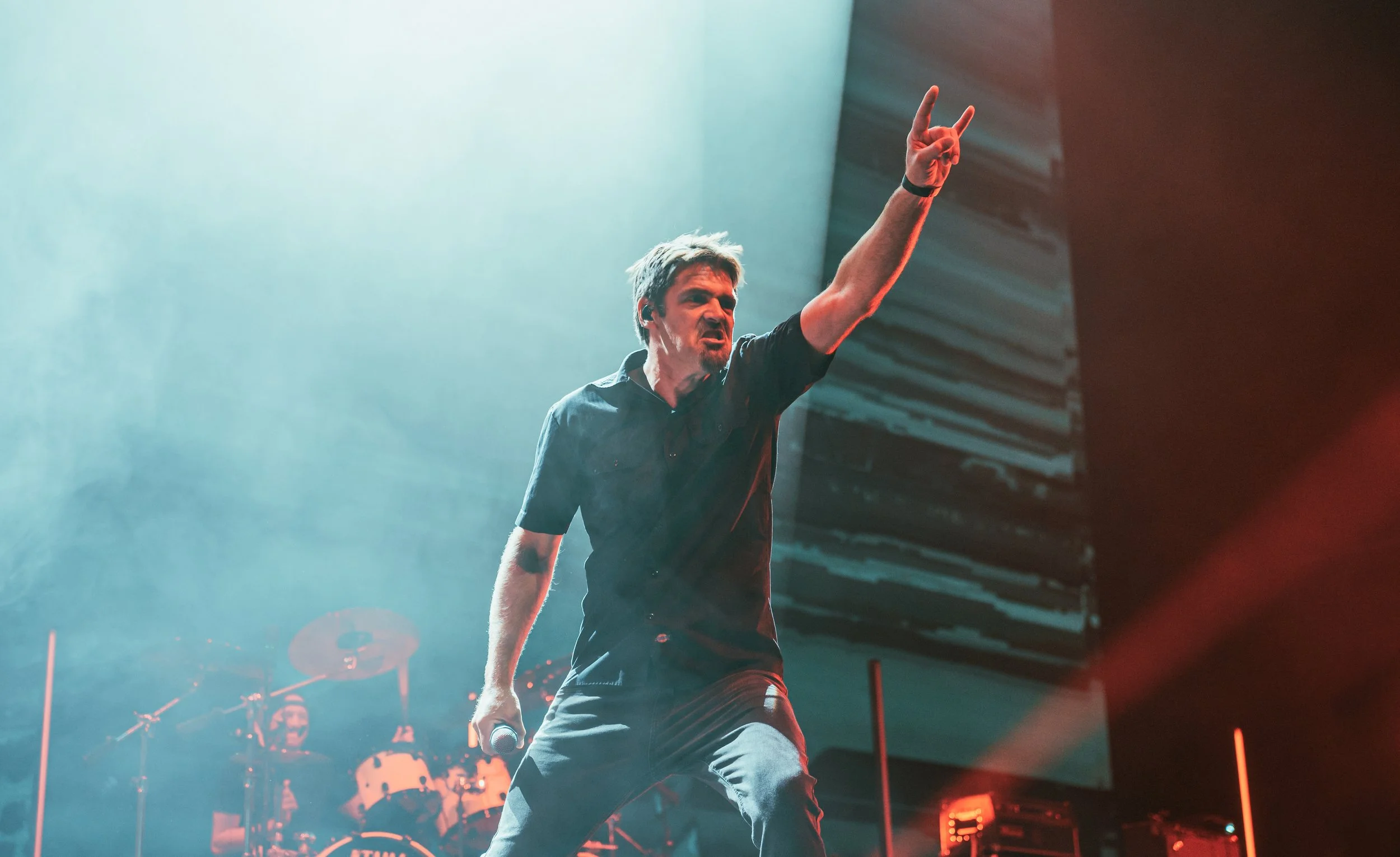 Male singer performing on stage with a microphone in his left hand, his right hand raised in victory, against a backdrop of stage lights and band equipment.