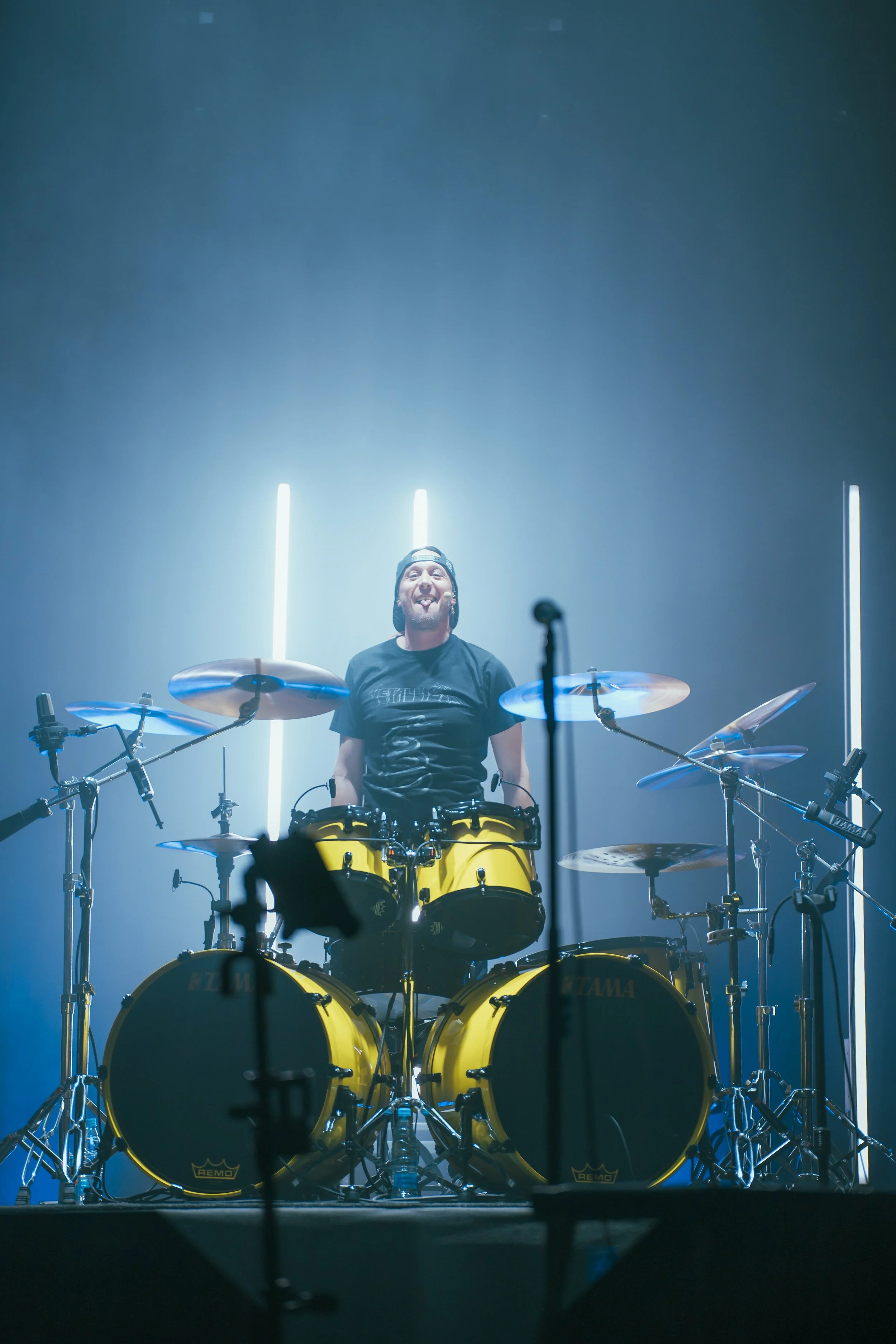 A musician standing behind a yellow drum set on stage, smiling, with stage lights and a dark background.