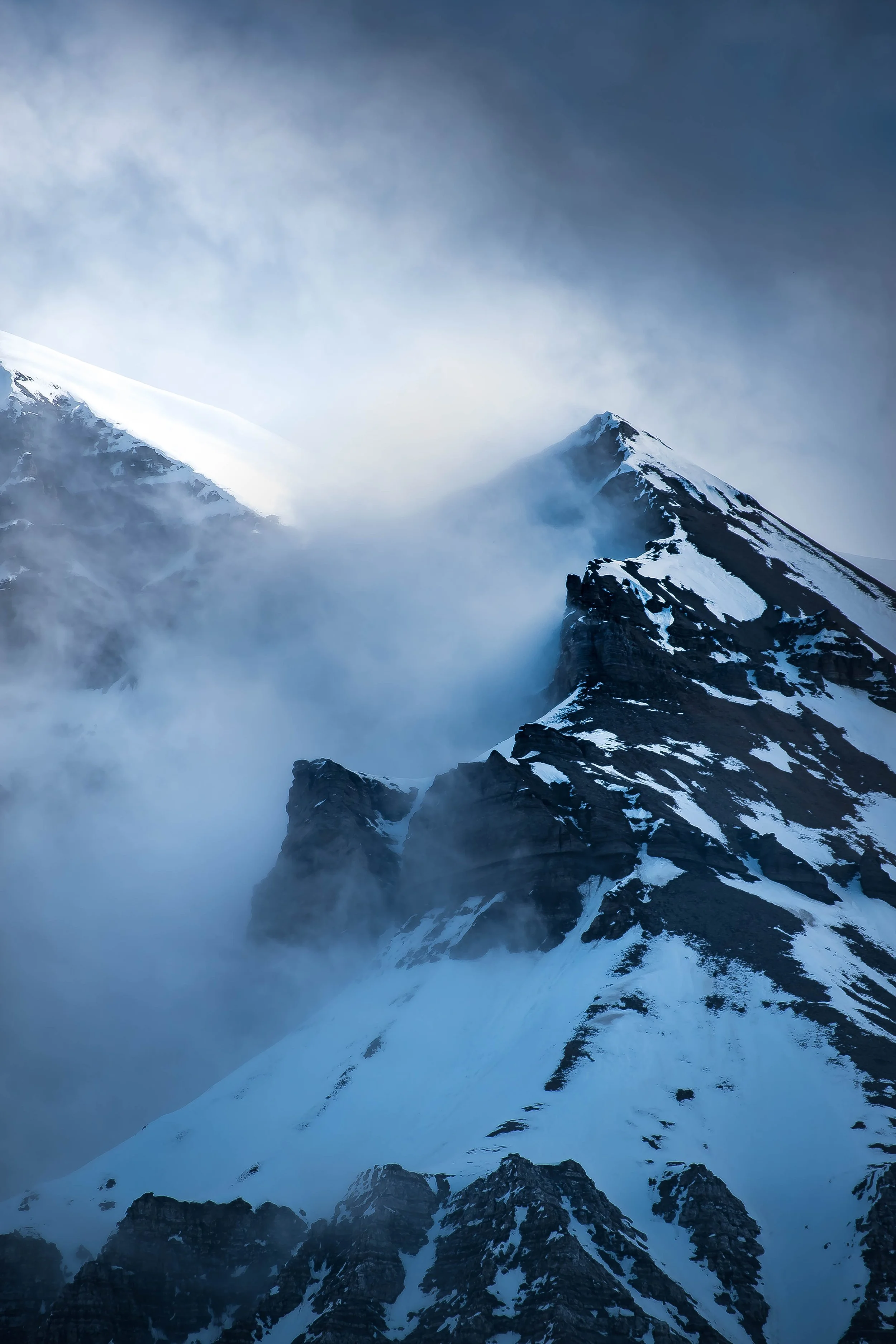 Snow-capped mountains with steep, rocky slopes and misty clouds surrounding the peaks.