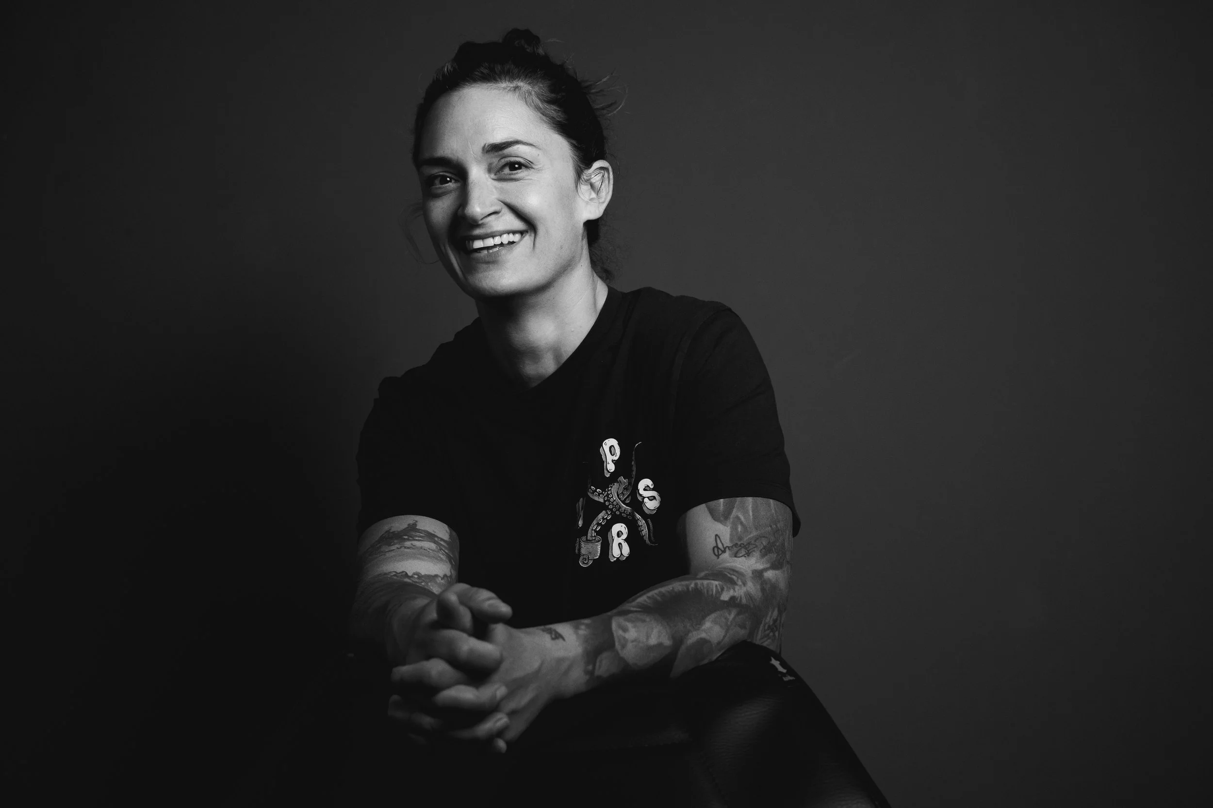 Black and white portrait of a smiling woman with tattoos, sitting with her hands clasped. She has dark hair tied back, wearing a black t-shirt with a design on the left side, and is positioned against a plain dark background.