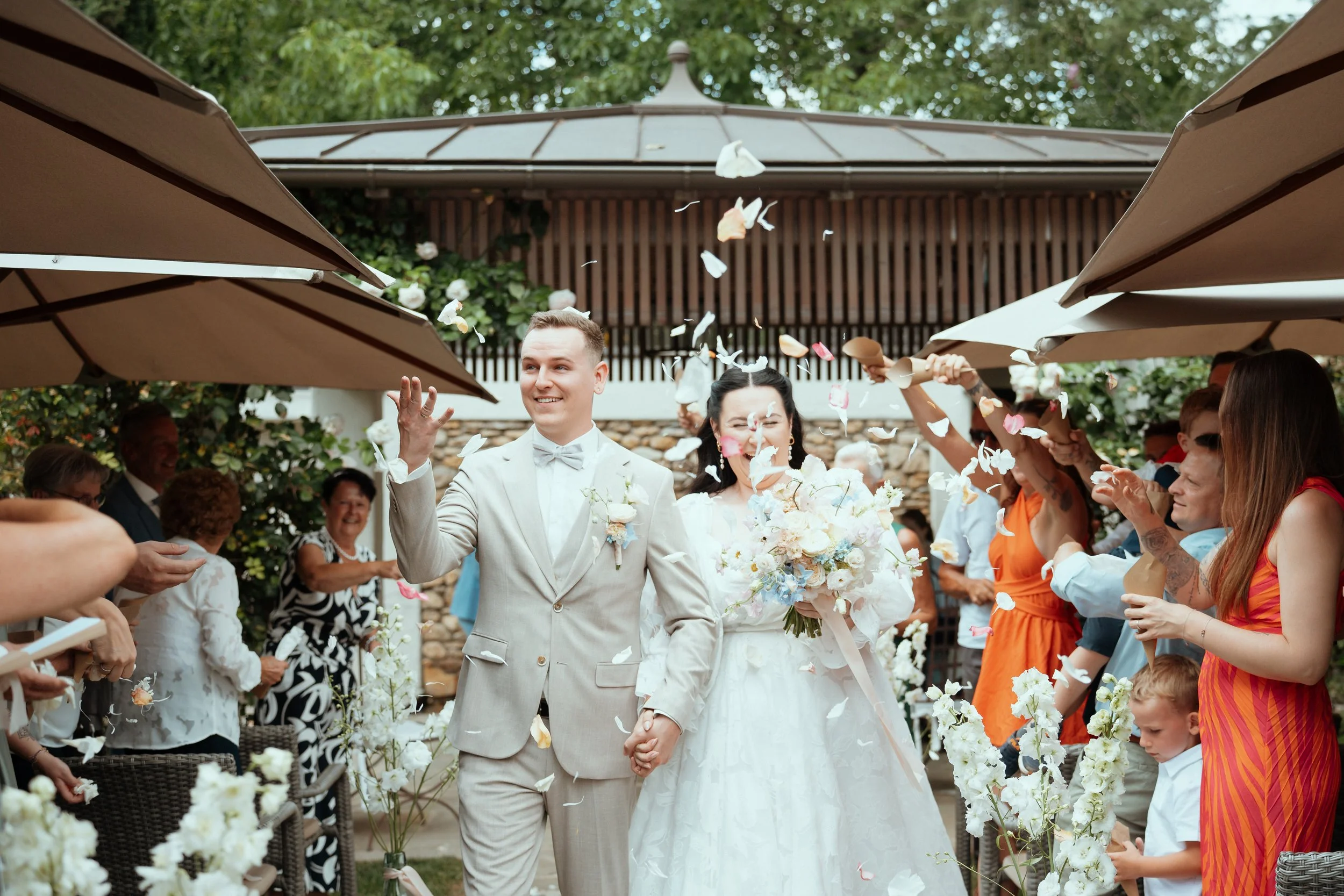 A wedding celebration outdoors with a bride and groom holding hands, the groom in a beige suit and bow tie, the bride in a white wedding dress holding a bouquet, and guests surrounding them throwing flower petals while smiling and taking photos under