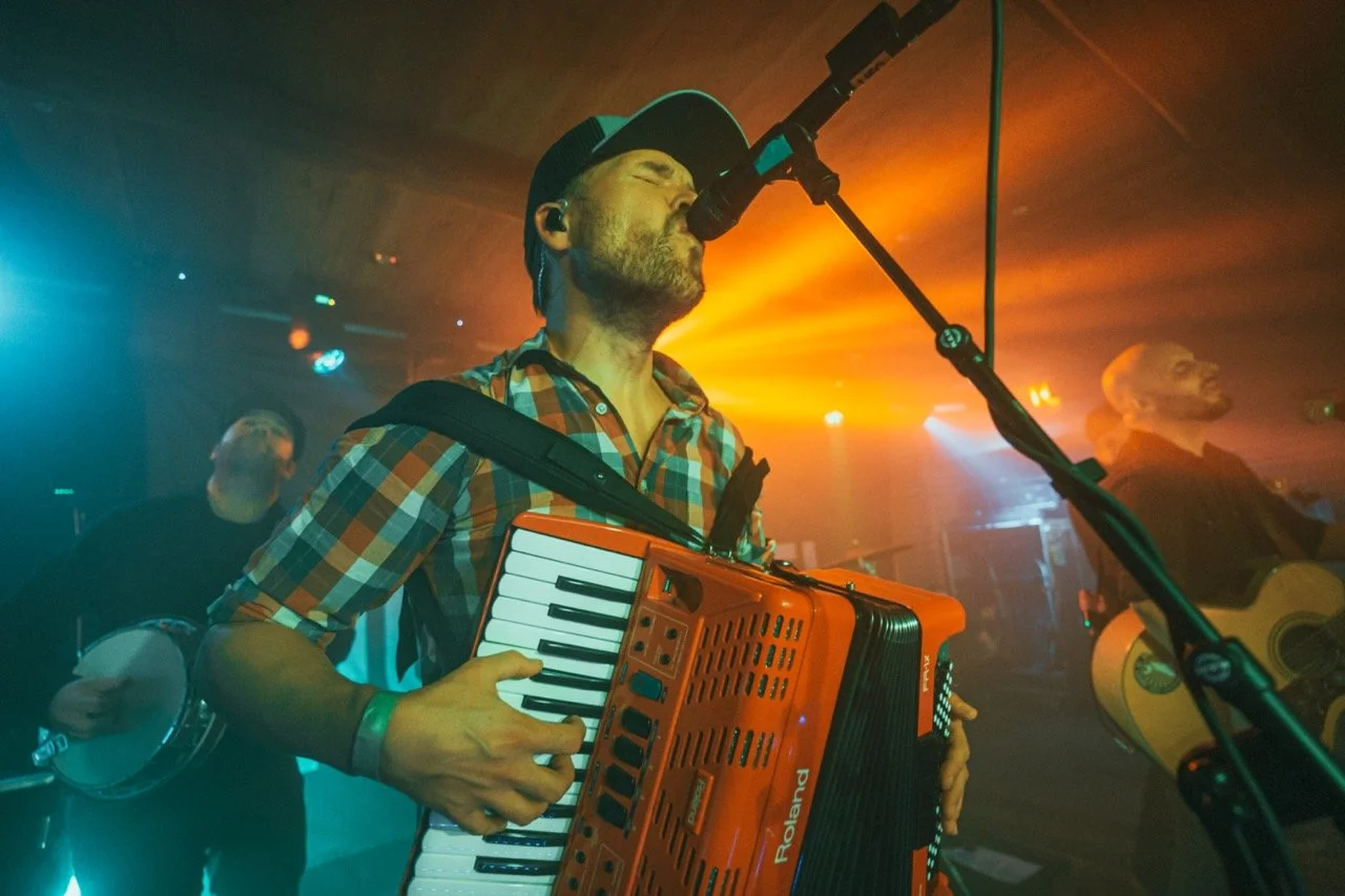 A man in a plaid shirt and baseball cap playing an orange accordion and singing into a microphone during a live music performance with band members in the background and colorful stage lighting.
