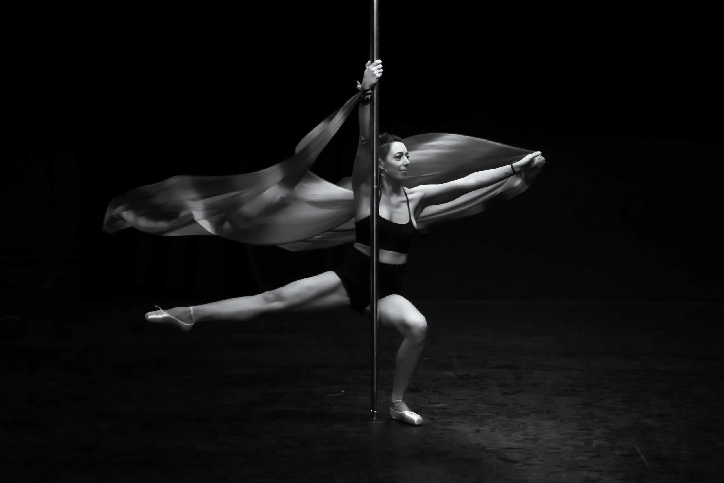 A female dancer performs a pose with a pole, wearing ballet attire, with flowing fabric behind her, in a dark studio.