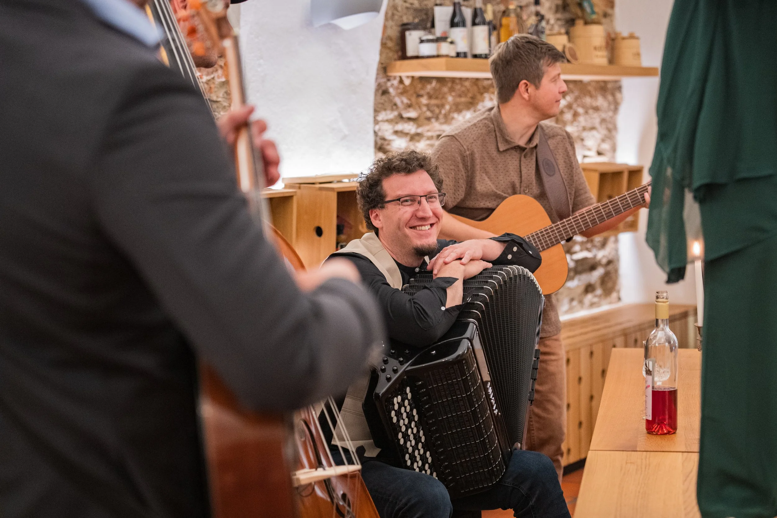 A man smiling and leaning on an accordion, sitting at a table with a bottle of red liquid. Behind him, two men are playing guitars in a cozy, rustic setting.
