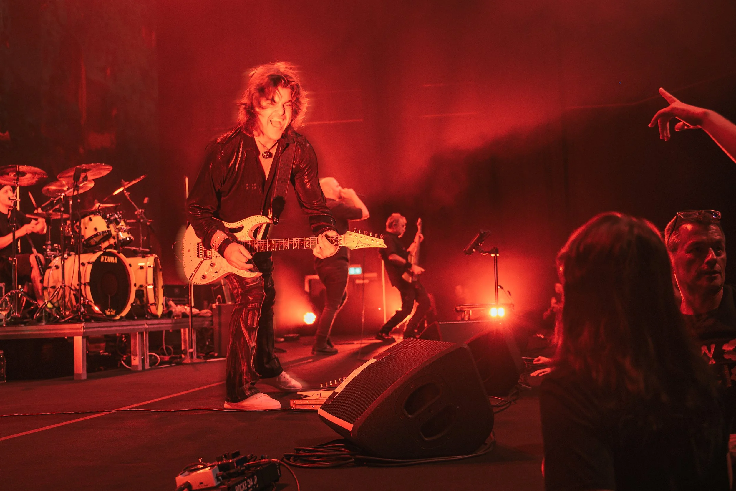 A rock band performing live on stage with red lighting, featuring a guitarist in the foreground intensely playing guitar, a drummer, and other band members playing instruments in the background, with audience members visible near the stage.