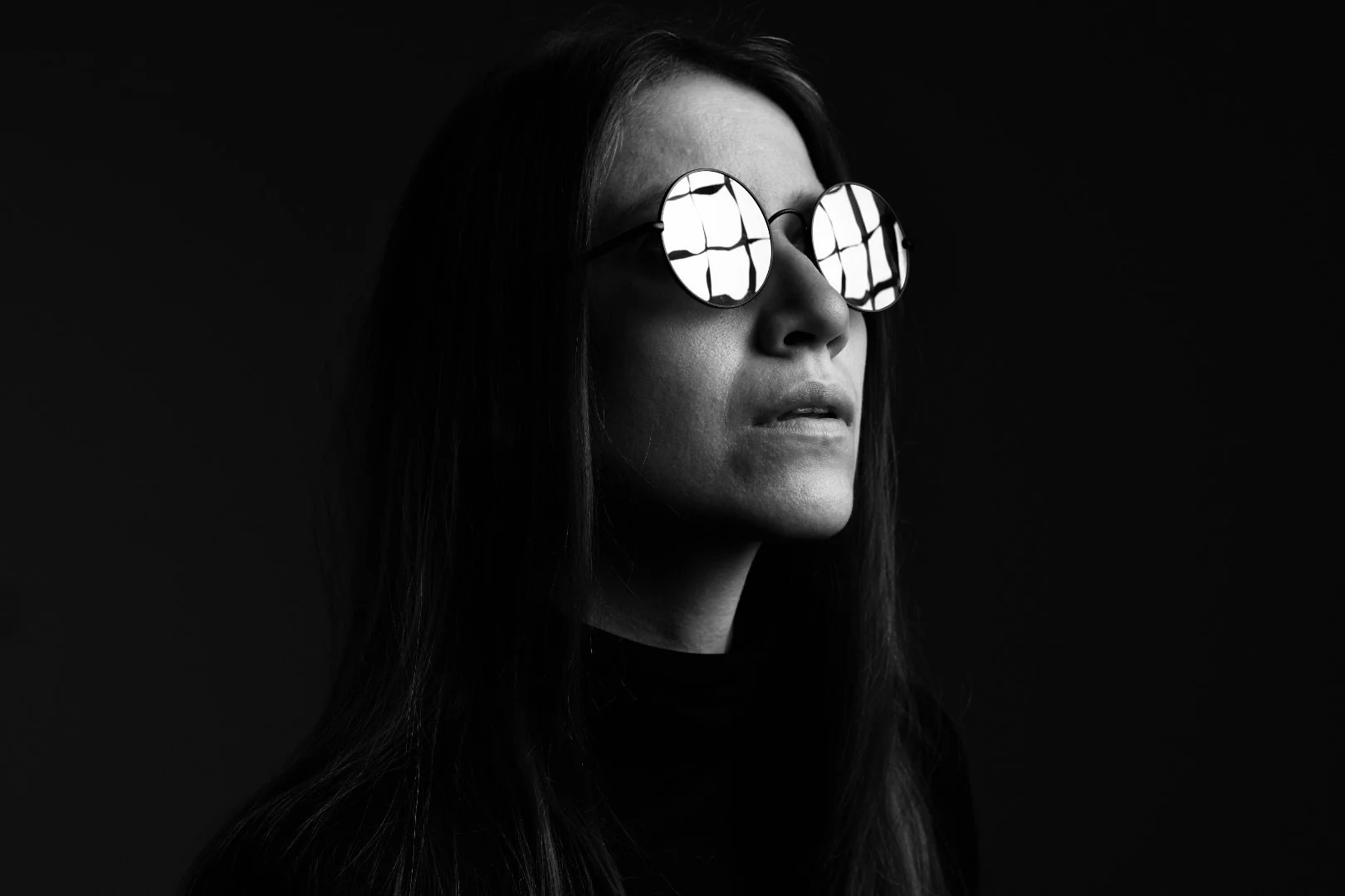 Black and white portrait of a woman wearing round sunglasses, with reflections of window panes in the lenses, against a dark background.
