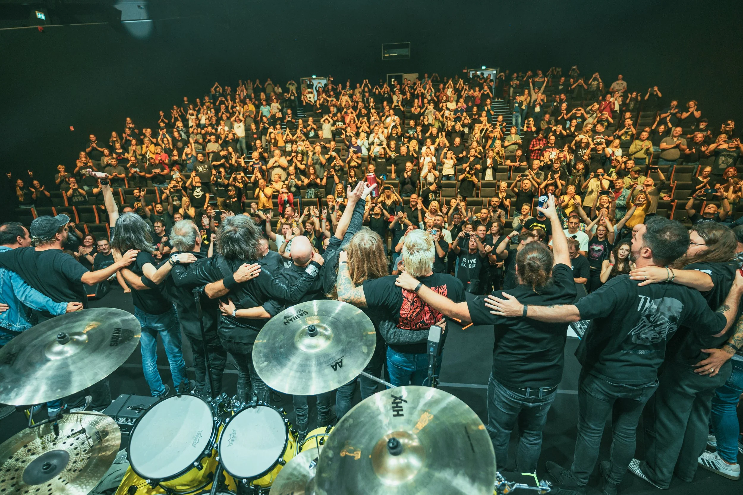 A large crowd at a concert or event, with many people standing and raising their hands, as the band on stage embraces each other at the front.