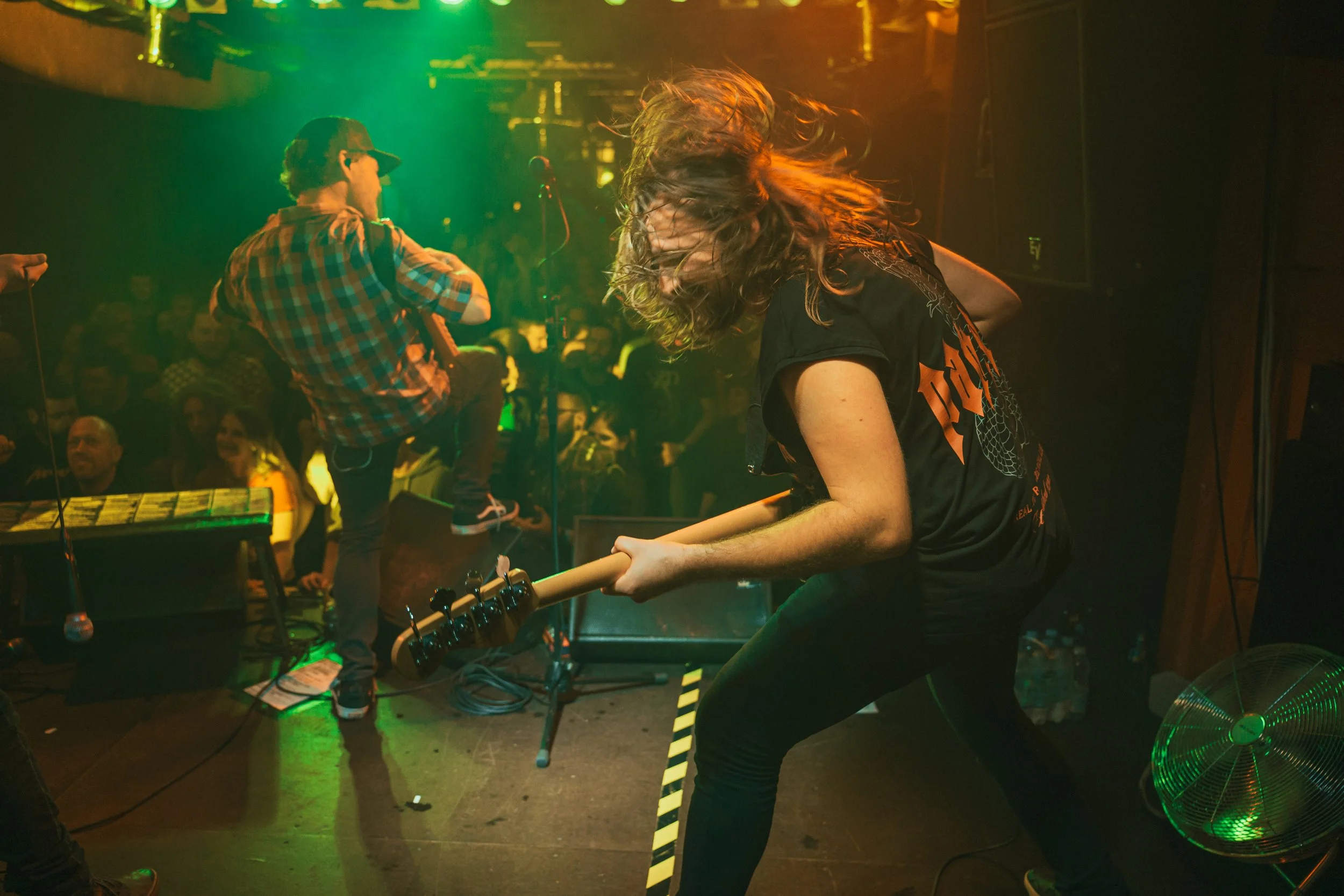 A live music performance with two musicians on stage, one playing guitar and the other on an electric guitar. The guitarist with long hair is intensely focused, and the audience is visible in the background under colorful stage lights.