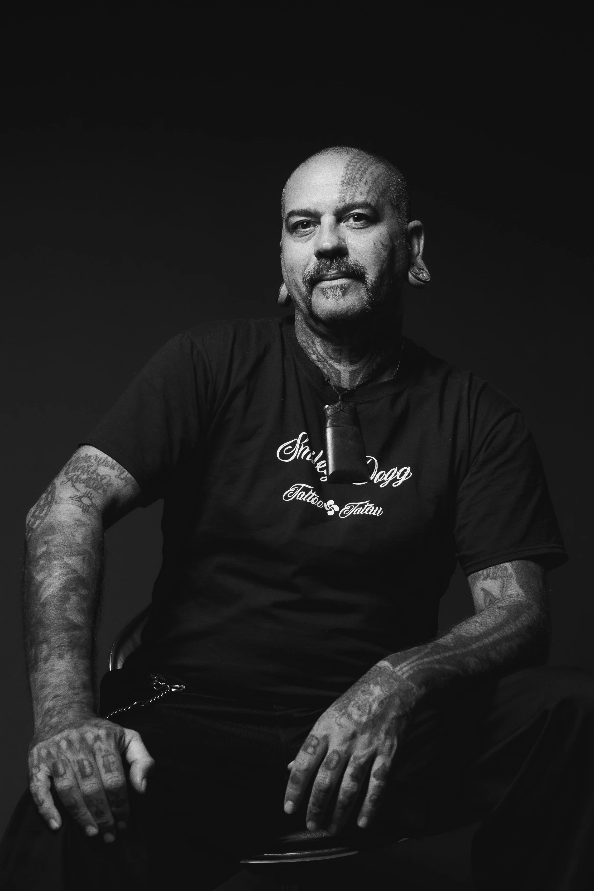 A tattooed man with a shaved head and earrings, sitting against a dark background.