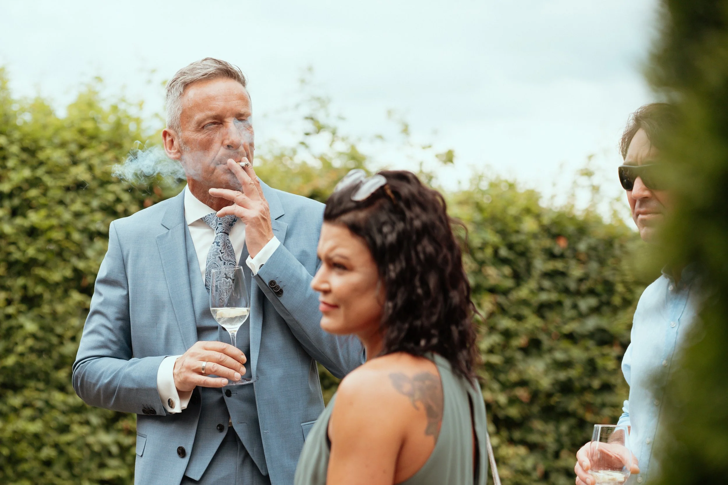 Three people at an outdoor gathering, one man in a blue suit smoking a cigarette and holding a glass of wine, a woman with dark curly hair and a tattoo on her shoulder, and another man wearing sunglasses and a light blue shirt, with greenery in the b