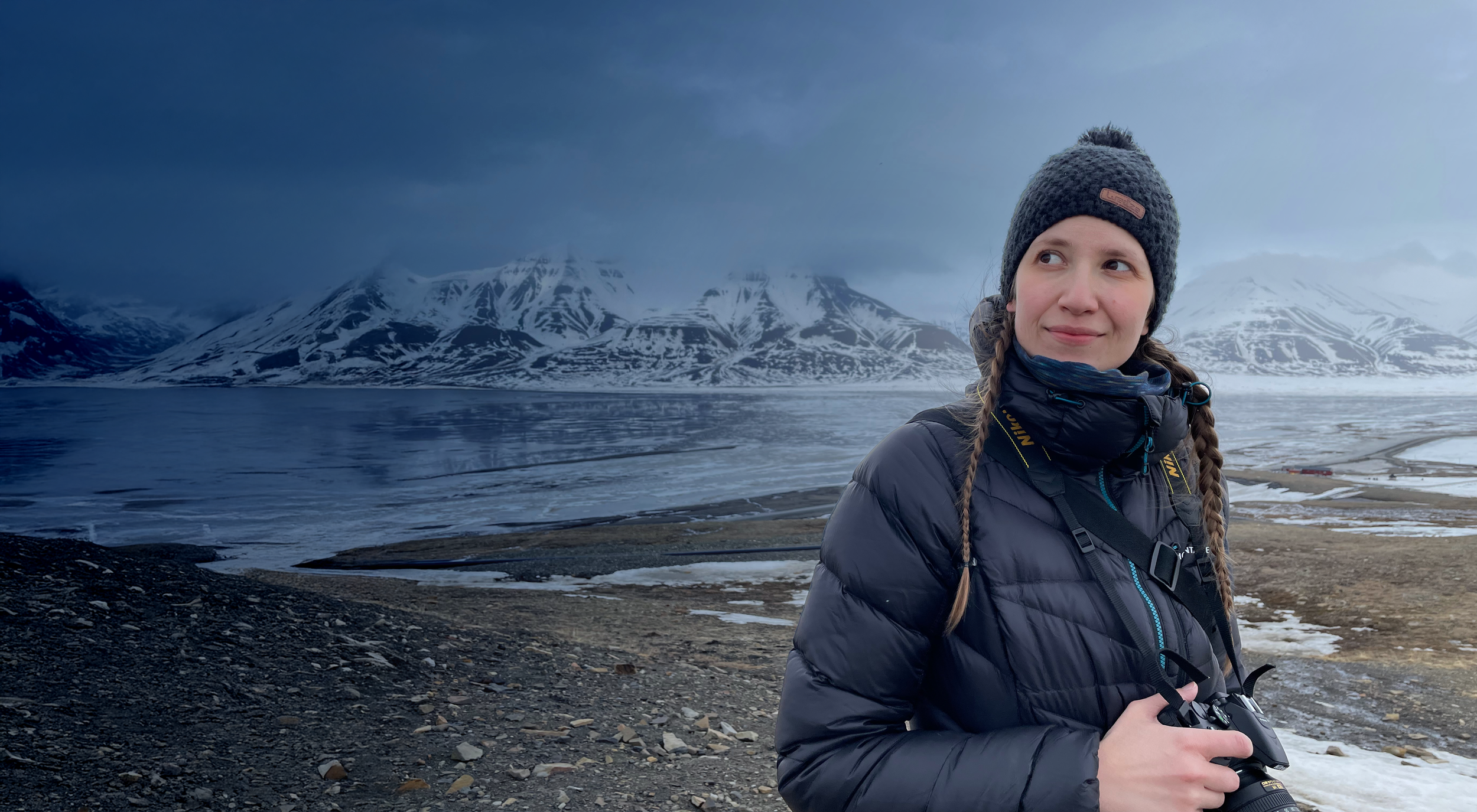A woman with braided hair, wearing a gray knit hat and a dark puffy jacket, stands on a rocky shoreline holding a camera with mountains and a lake in the background under cloudy skies.