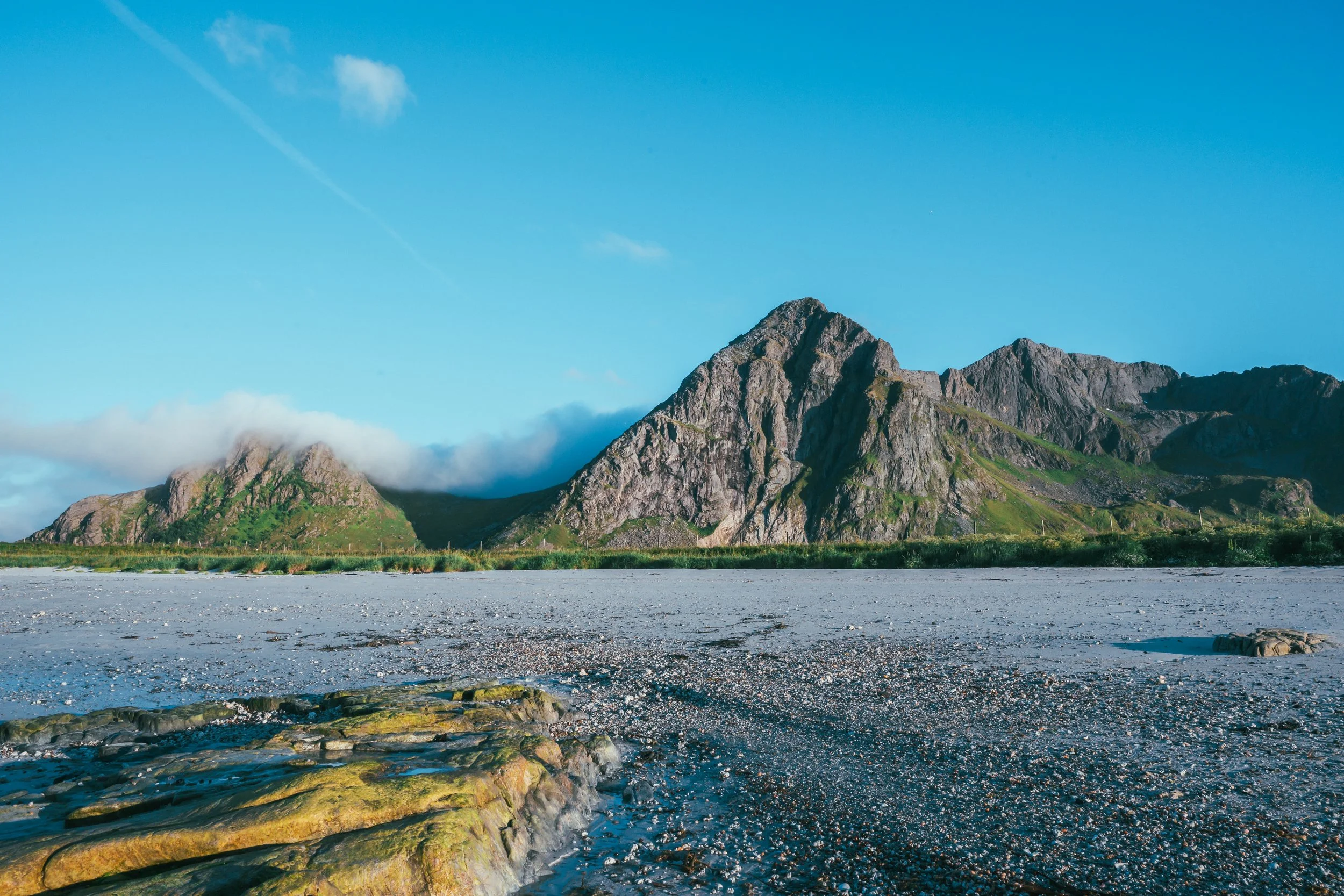 Mountains with a clear blue sky, some clouds, and a body of water with rocks in the foreground.