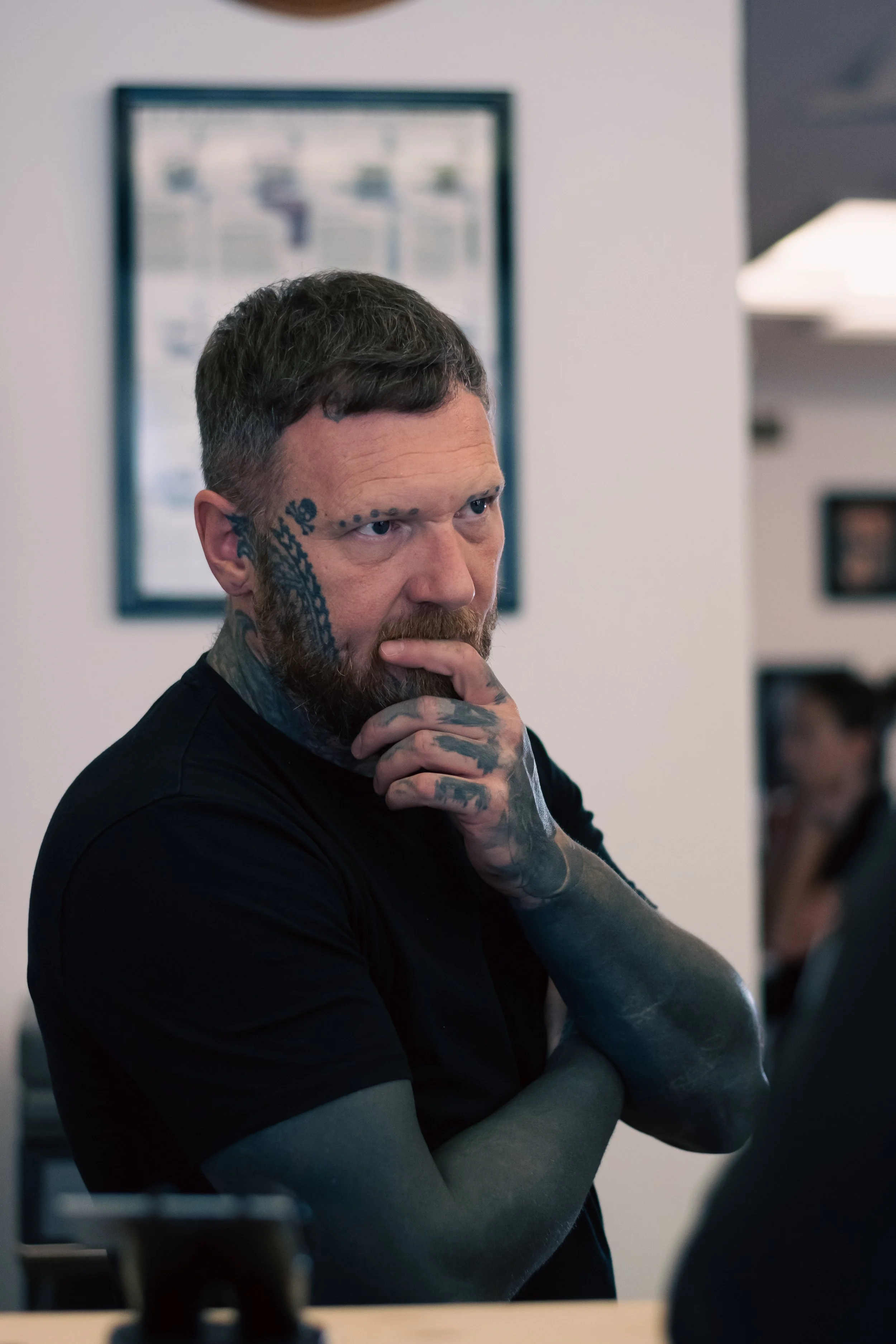 A man with tattoos on his face, arms, and hands, wearing a black shirt, standing indoors with a thoughtful expression, hand on chin